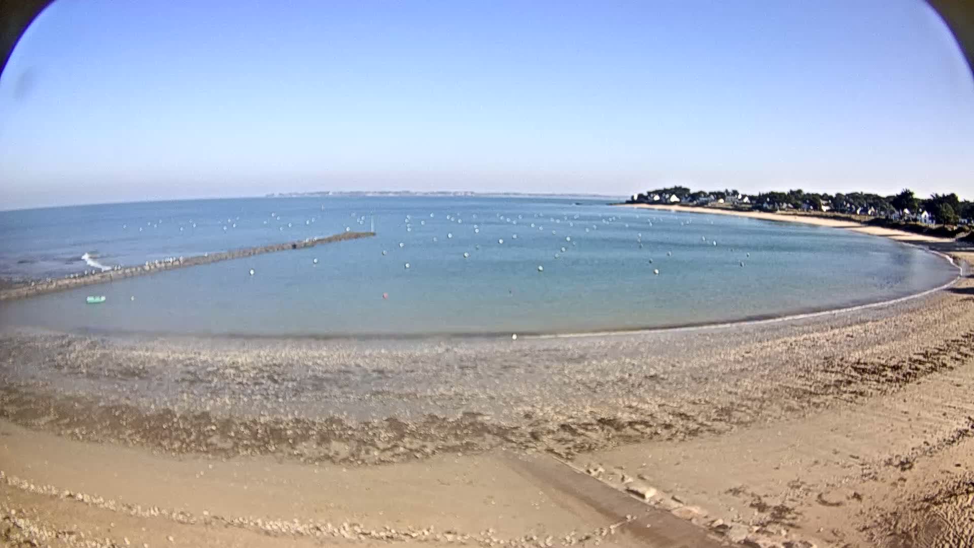 A sandy beach curves around a calm, light-blue bay dotted with buoys under a clear blue sky.