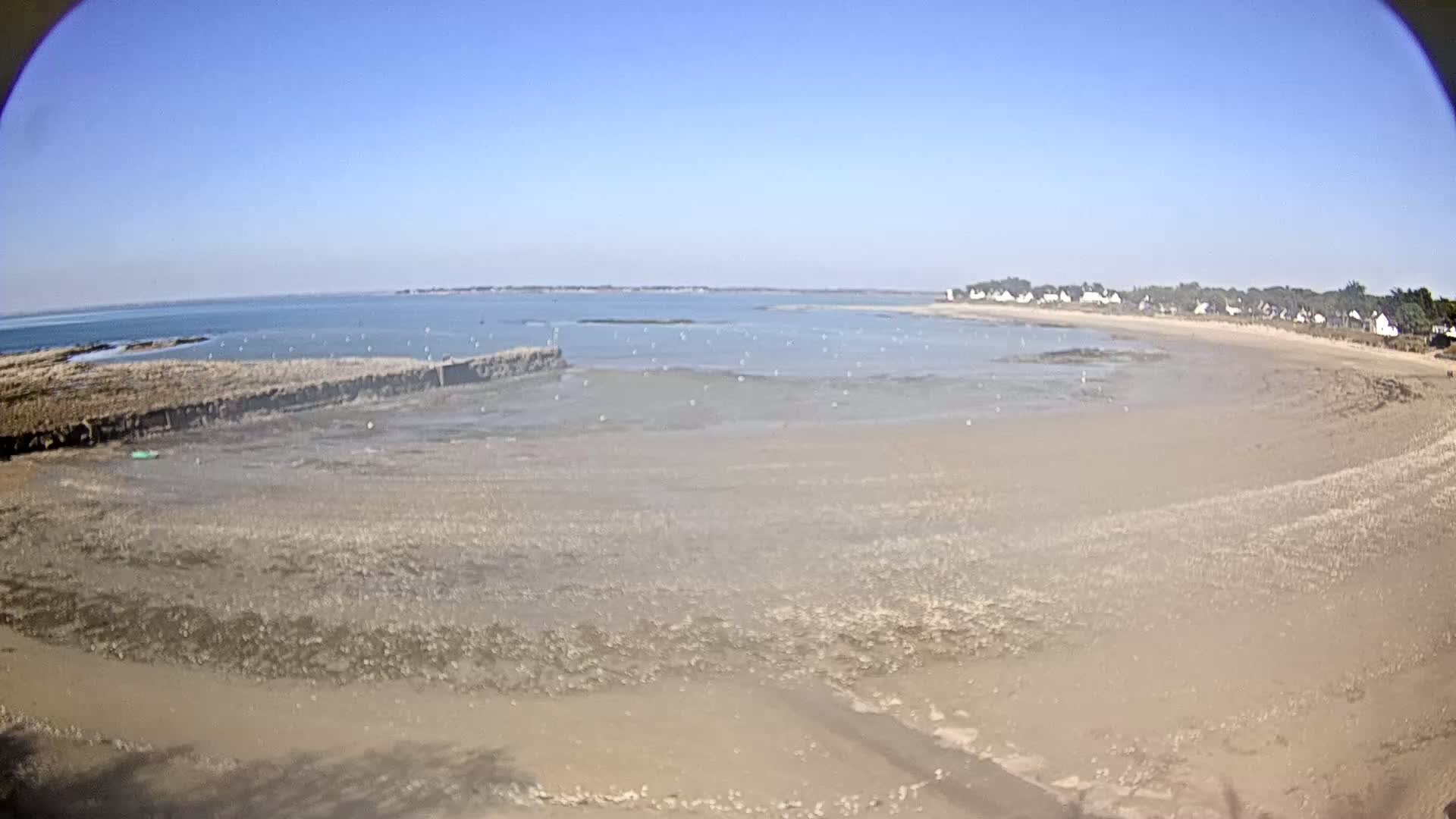 A wide sandy beach stretches along a calm bay under a clear blue sky, with a small village visible in the distance.