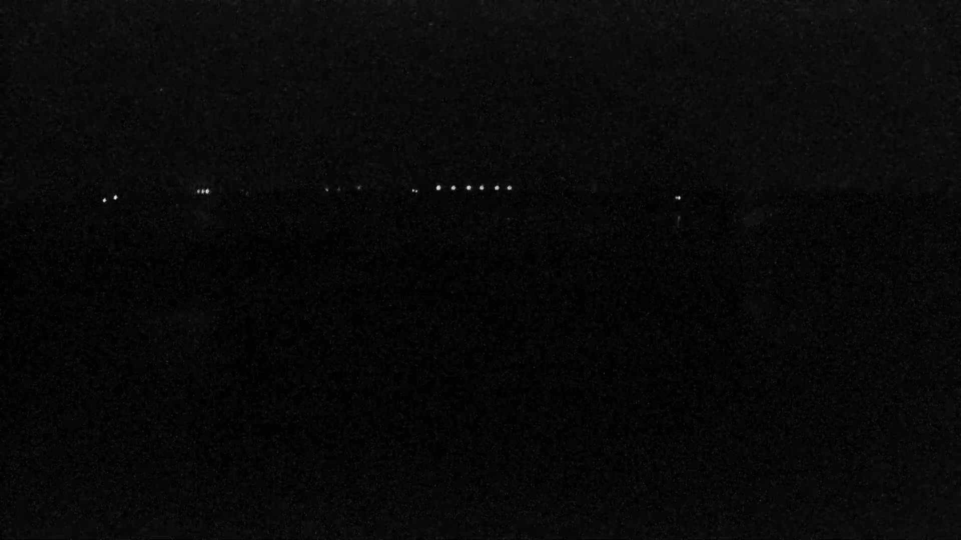 A dark, nighttime scene shows a line of faint lights in the distance.