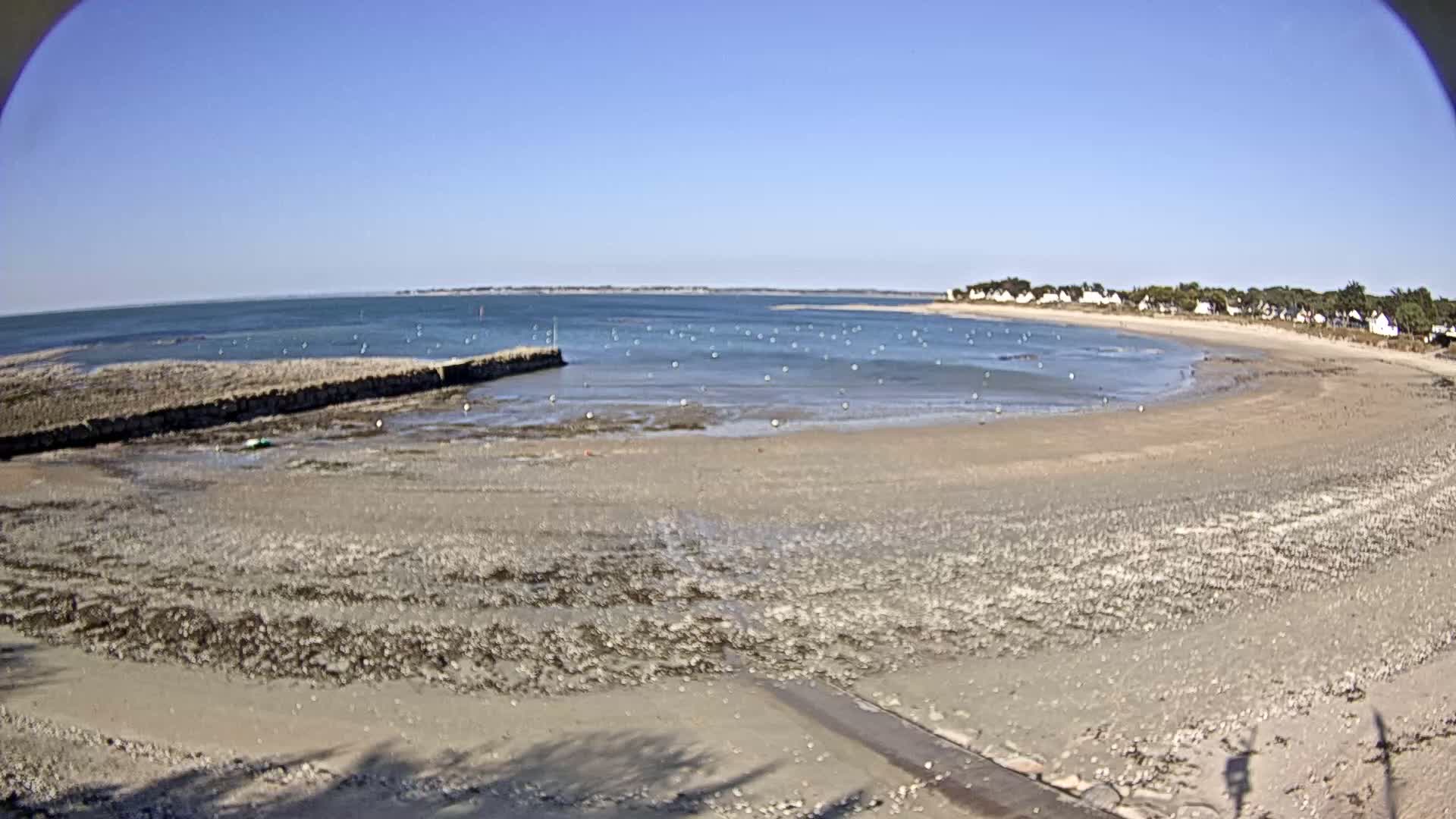 A mostly sunny day reveals a wide, sandy beach at low tide, with calm water, a stone jetty, and a line of houses visible in the distance.