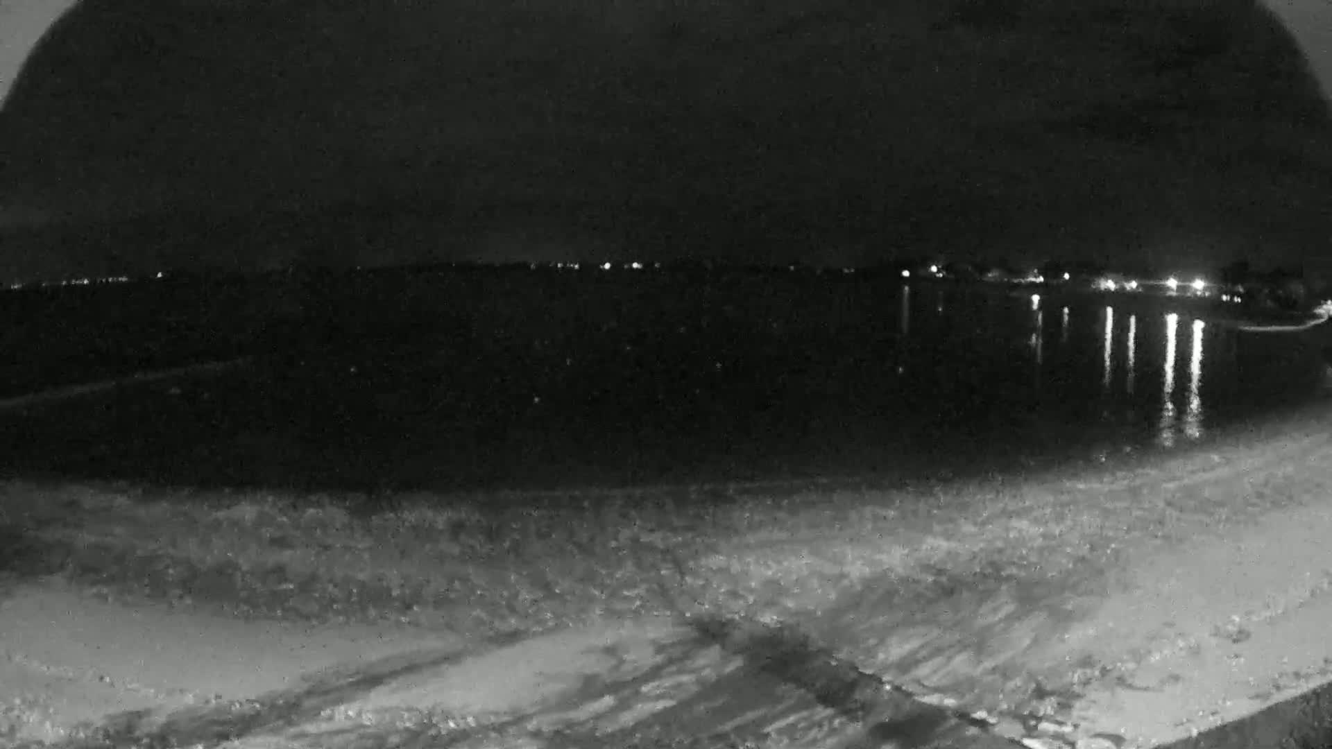 A nighttime, low-light view shows a dark body of water reflecting distant lights, with a sandy shore in the foreground.