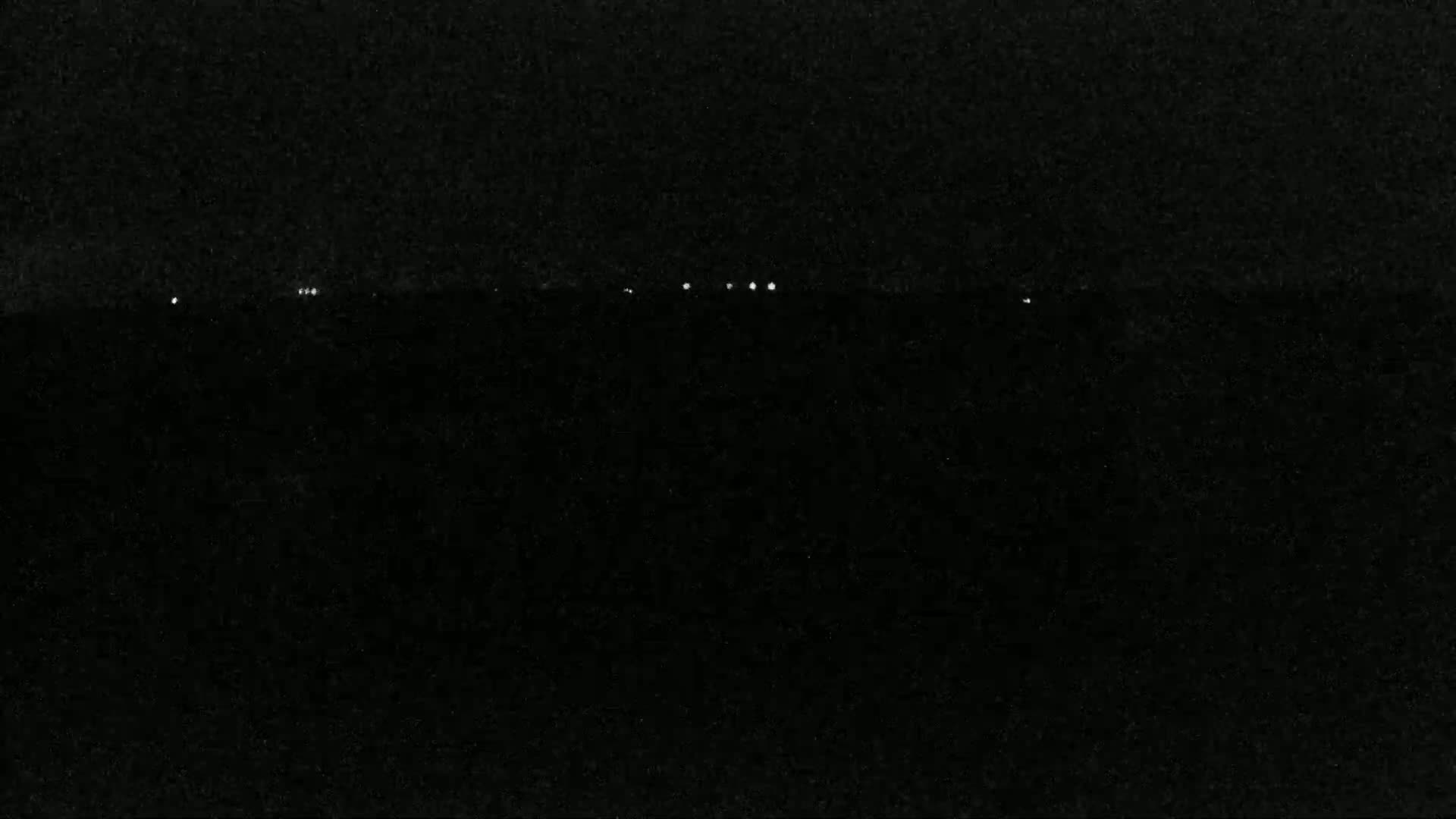 A nighttime image shows a dark, seemingly flat landscape with a few faint, distant lights on the horizon.