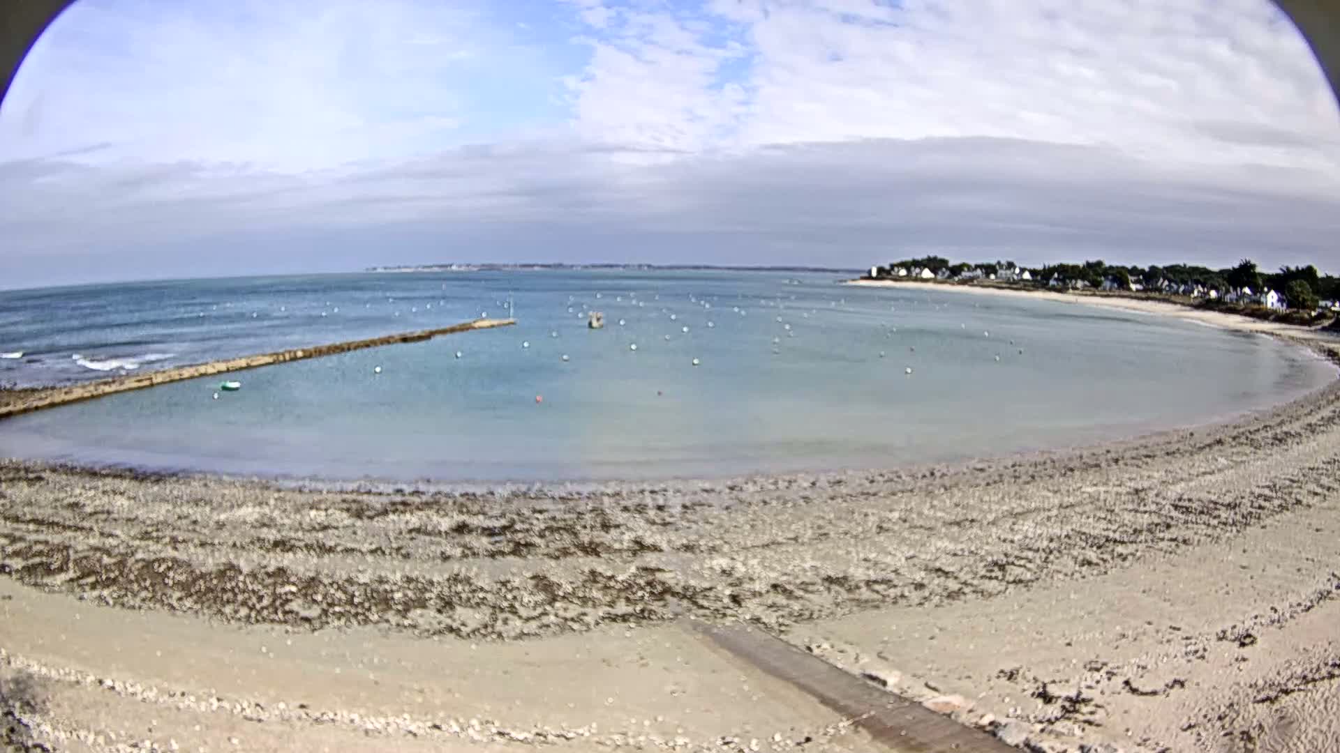 A calm, partly cloudy day reveals a sandy beach curving around a calm bay with a stone jetty, numerous buoys in the water, and a small village in the distance.