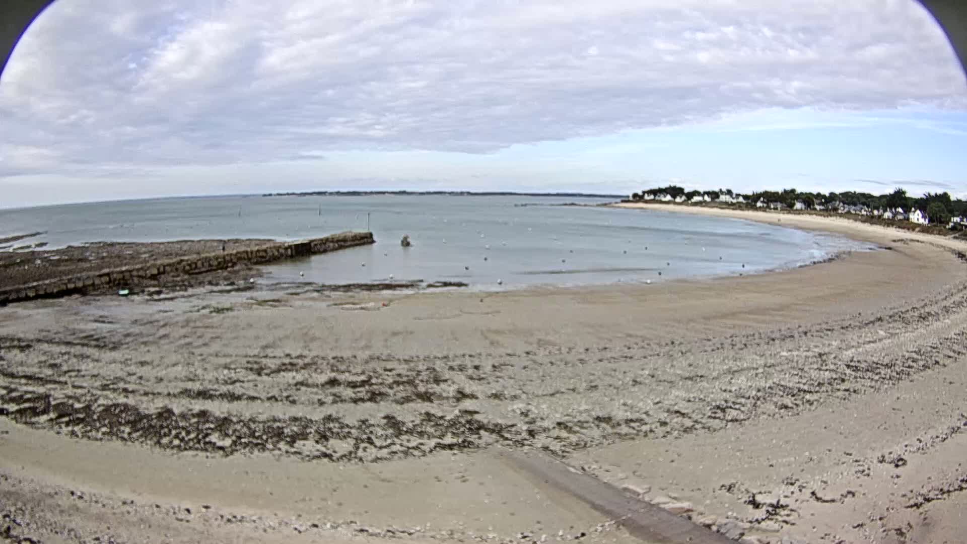 A wide, sandy beach curves around a calm bay under a partly cloudy sky, with a small stone jetty visible in the foreground.