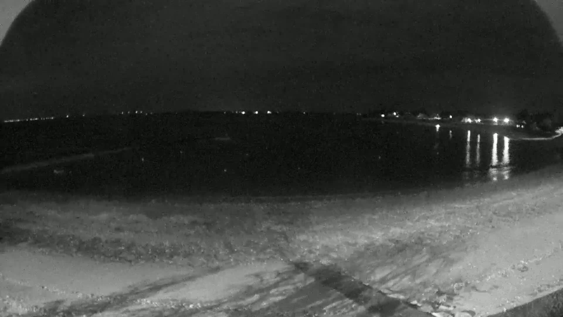 A dark, nighttime beach scene shows calm water reflecting distant lights, under a mostly clear sky.
