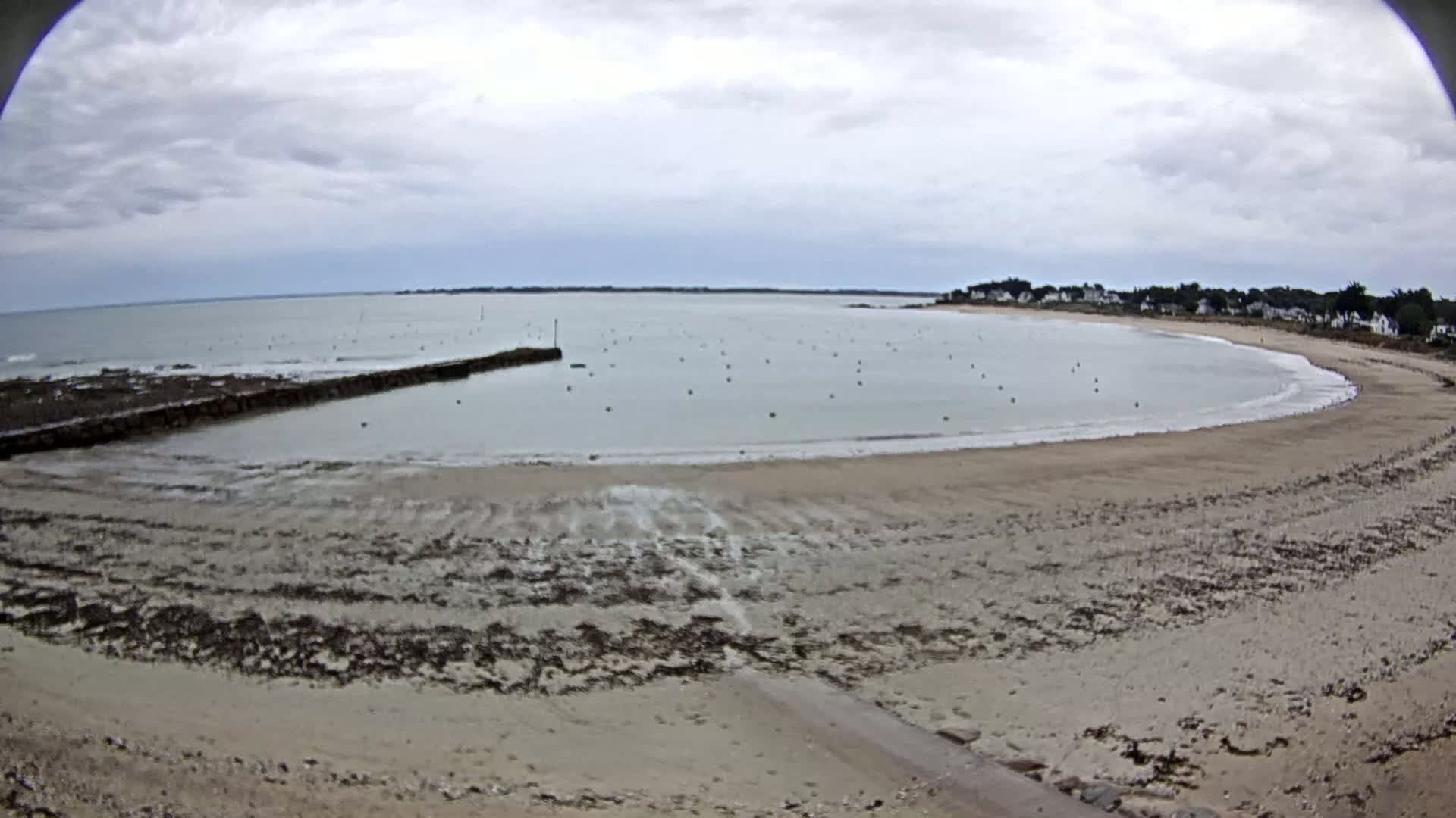 A sandy beach curves around a calm bay under an overcast sky, with a low stone wall separating a small harbor area.