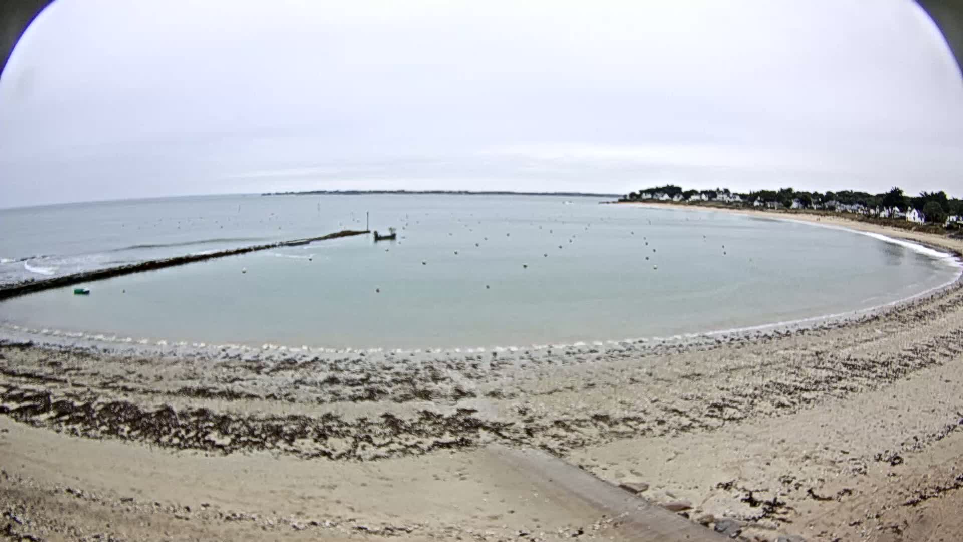 A calm, overcast day shows a sandy beach curving around a calm bay with a small boat and a few buoys visible in the water.