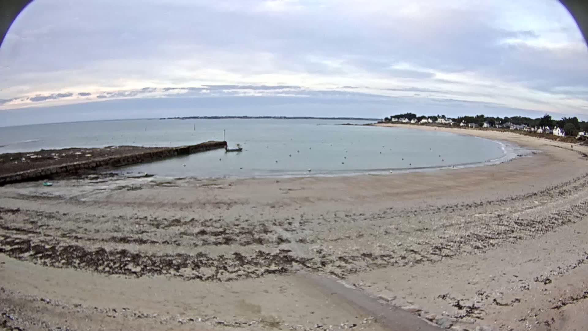 A mostly cloudy day reveals a sandy beach curving around a calm bay with a small boat and a stone jetty, near a line of houses.