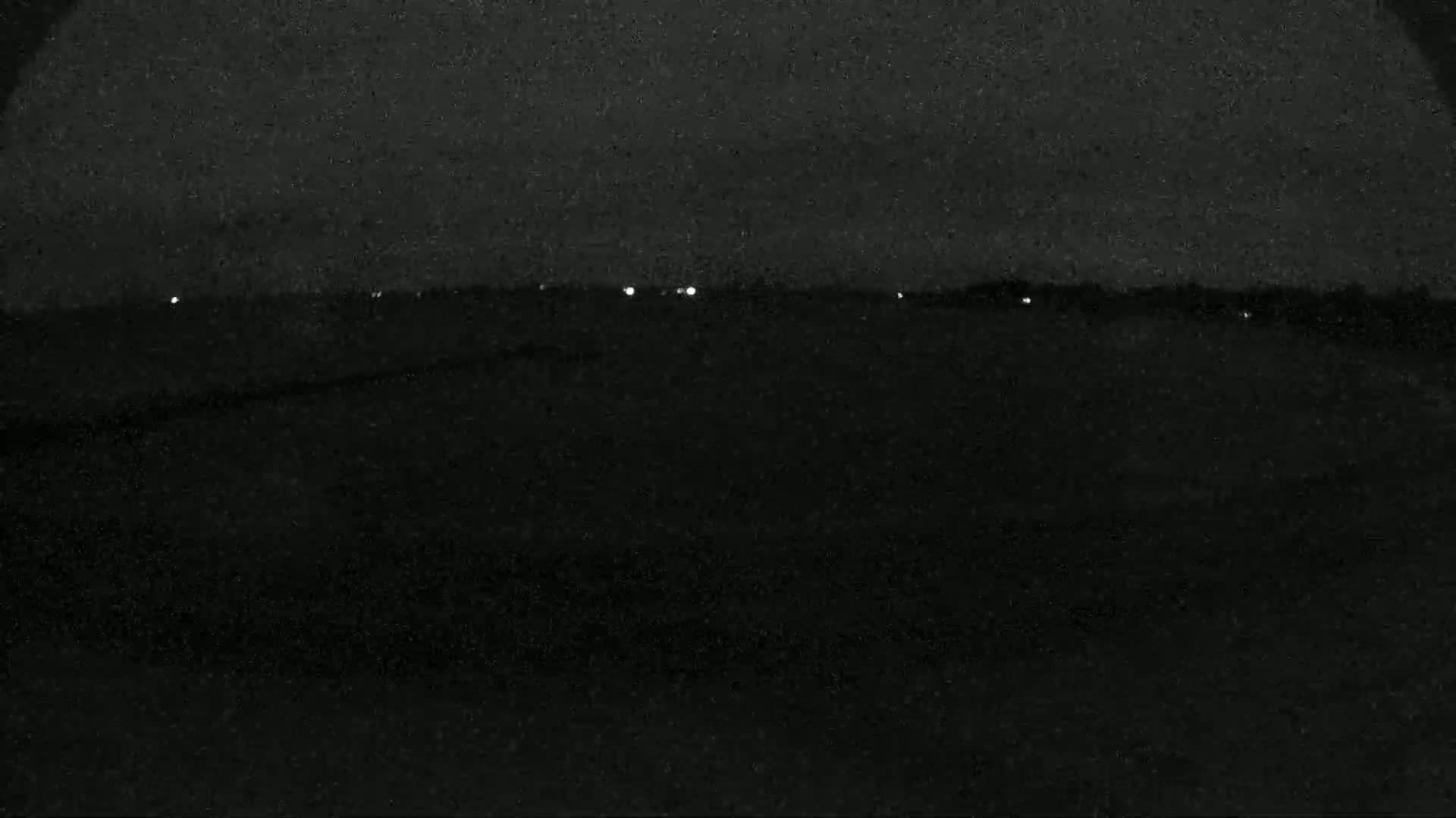 A nighttime, low-light view of a dark, flat landscape with a few distant lights visible on the horizon.