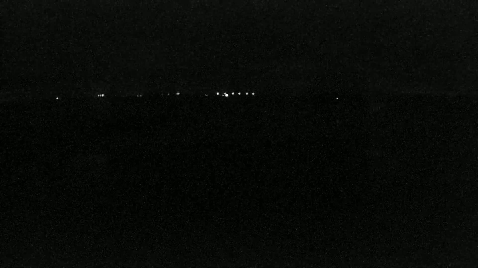 A nighttime, dark scene shows a line of small, distant white lights against a black background.