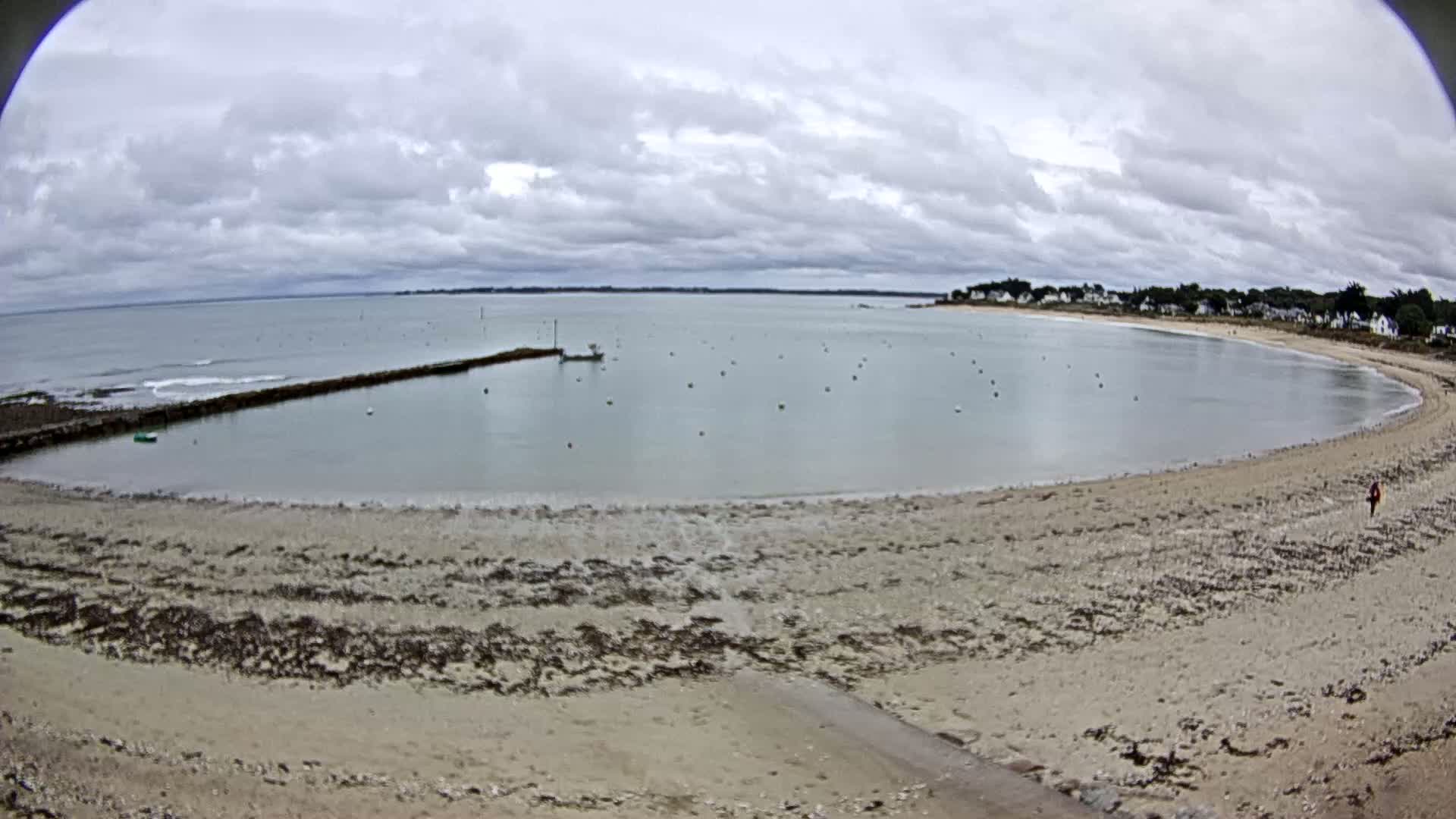 A sandy beach curves around a calm bay under a cloudy sky, with a small harbor and a few boats visible in the distance.