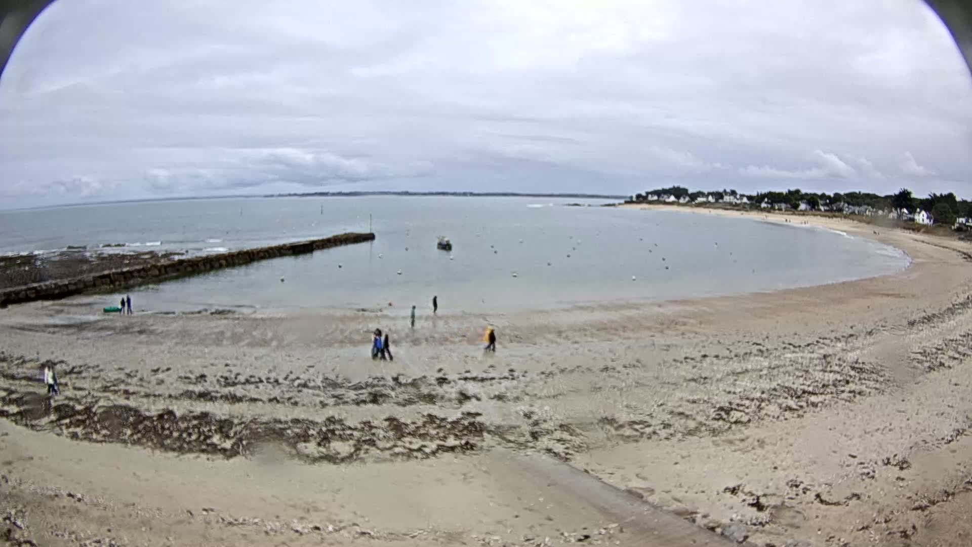 A mostly cloudy day reveals a sandy beach curving around a calm bay with several people walking and a small boat on the water.