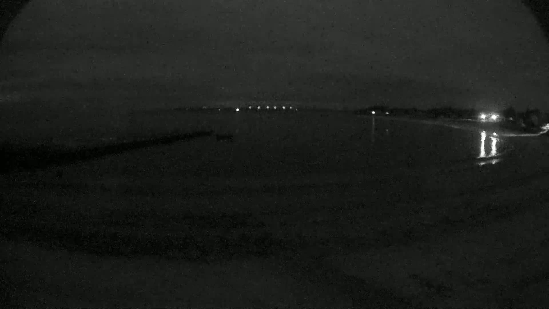 A nighttime, low-light view of a dark, seemingly calm body of water with a shoreline featuring faint lights in the distance and one or two indistinct figures near the water's edge.