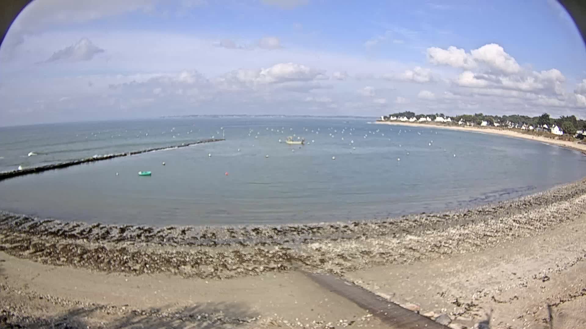 A partly cloudy day shows a calm bay with a small boat, a rocky beach, and a distant shoreline with houses under a mostly clear sky.