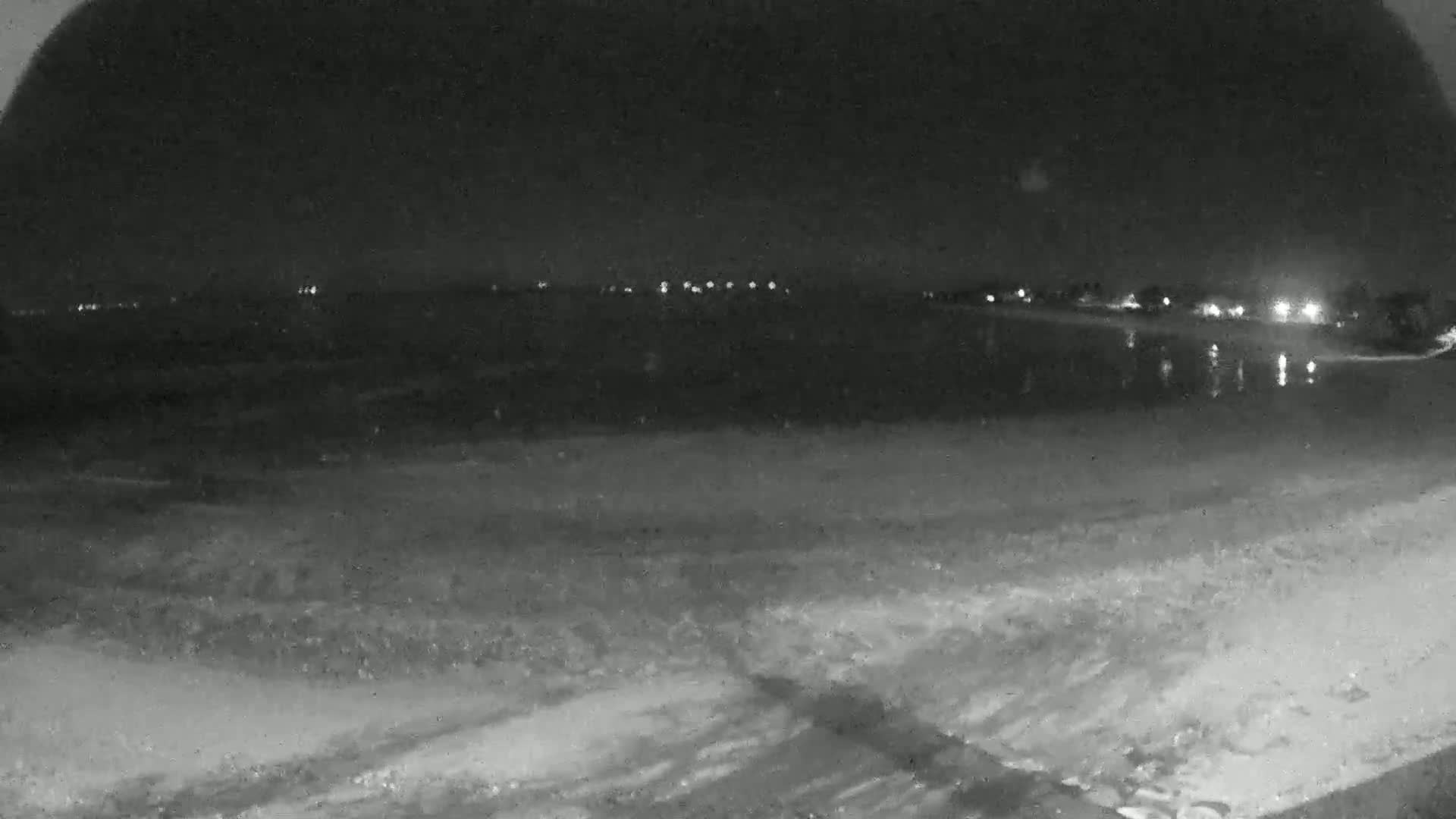 A nighttime, low-light view of a dark beach and calm water with distant lights visible on the horizon.