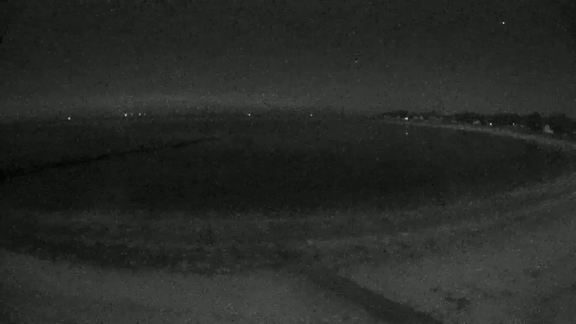A nighttime, low-light view shows a dark, seemingly flat landscape with a few distant lights visible on the horizon under a clear, starry sky.
