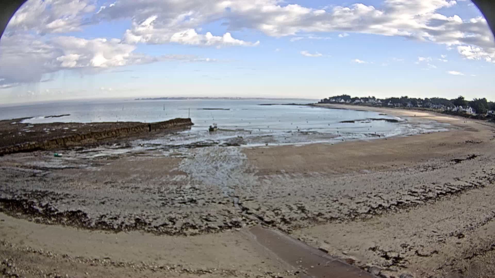 A mostly sunny day shows a wide sandy beach at low tide, with a small boat visible near a low rock wall and a small village in the distance.
