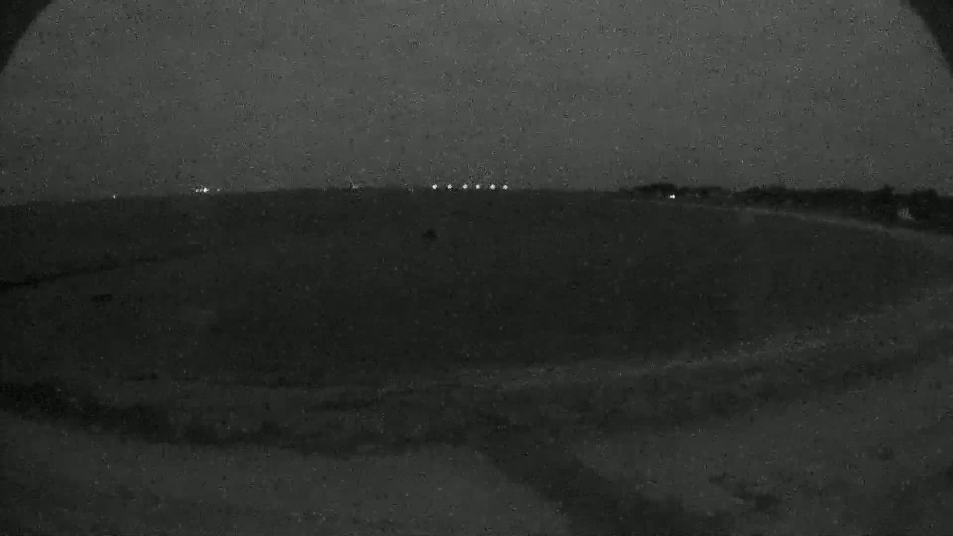 A dark, nighttime scene shows a flat expanse of land with a line of distant lights on the horizon under an overcast sky.