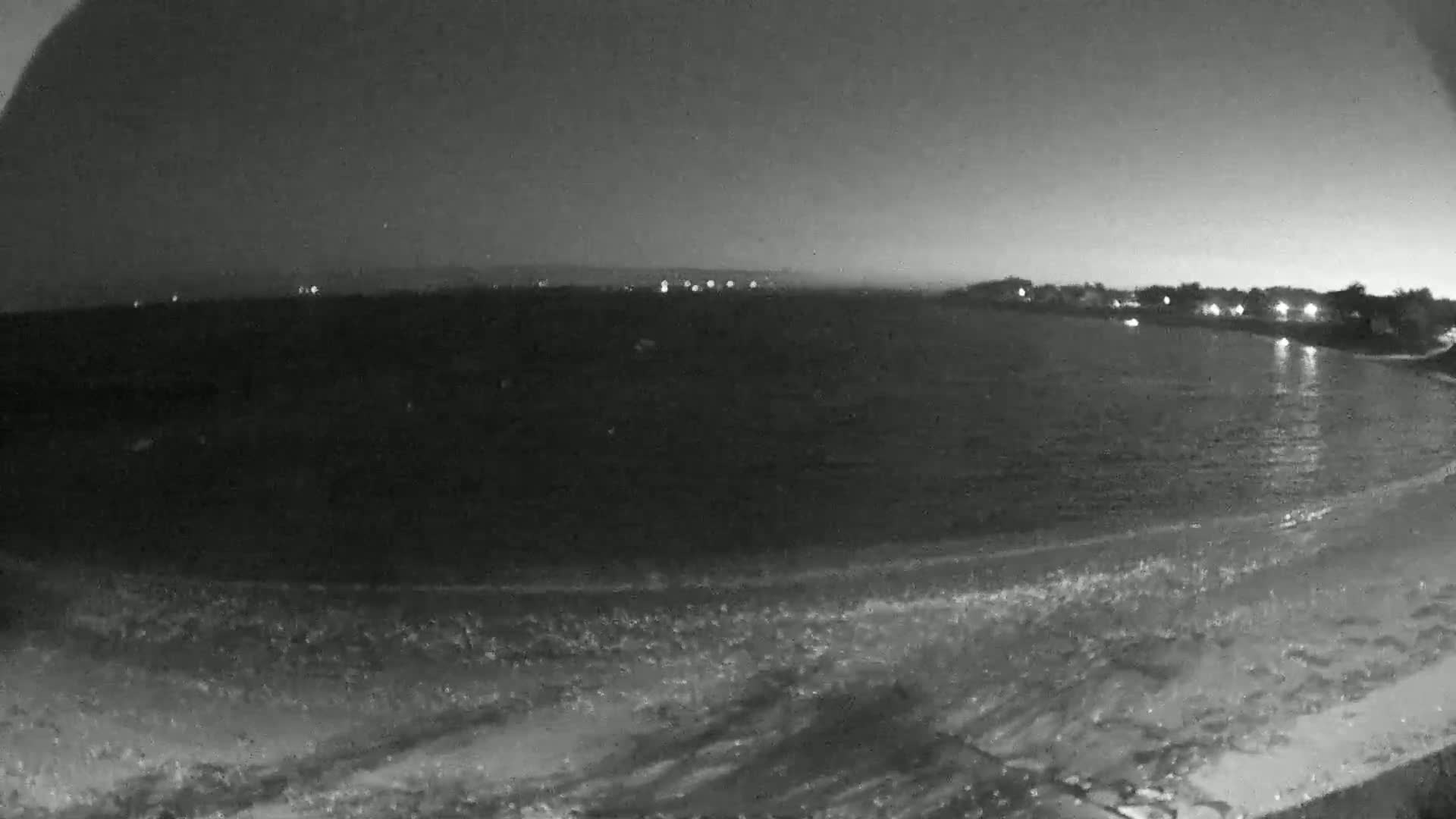 A nighttime, grayscale view of a calm sea lapping a sandy shore, with dimly lit land visible in the distance.