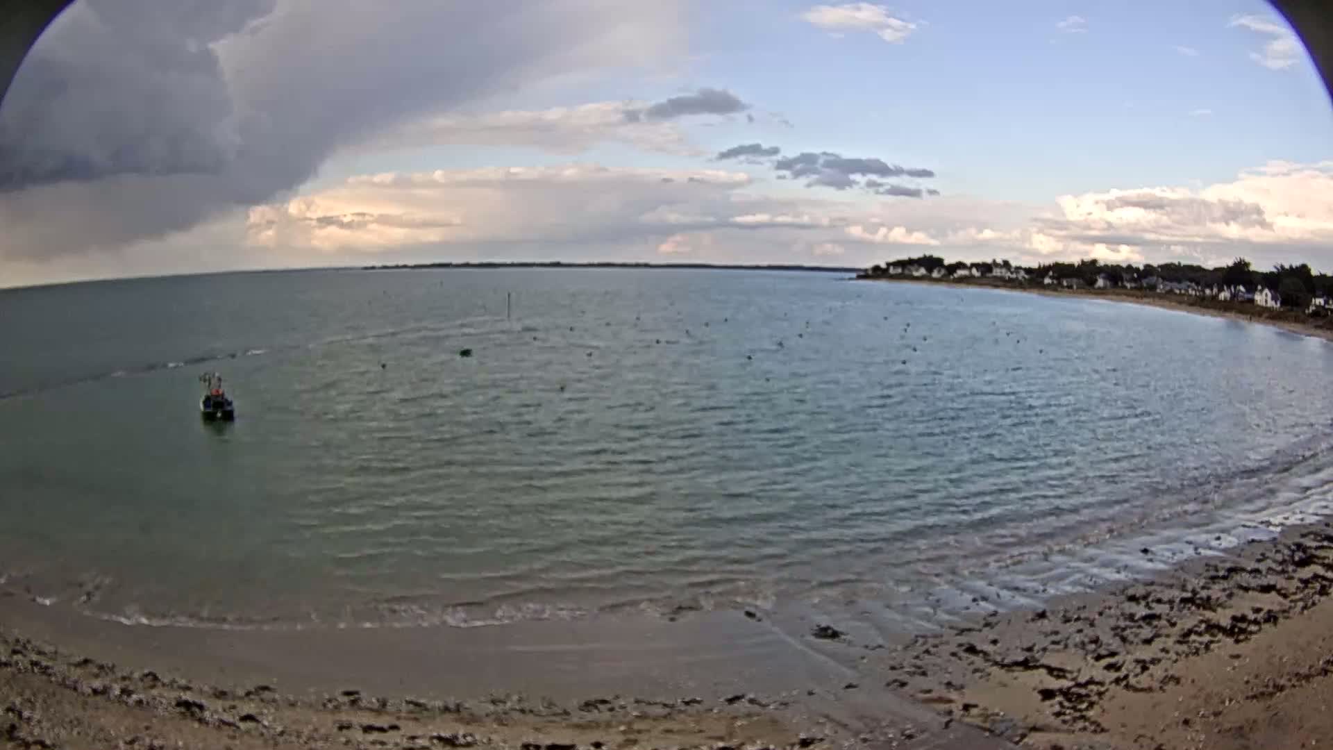 A calm bay with a sandy beach and a small boat is visible under a partly cloudy sky with some storm clouds.