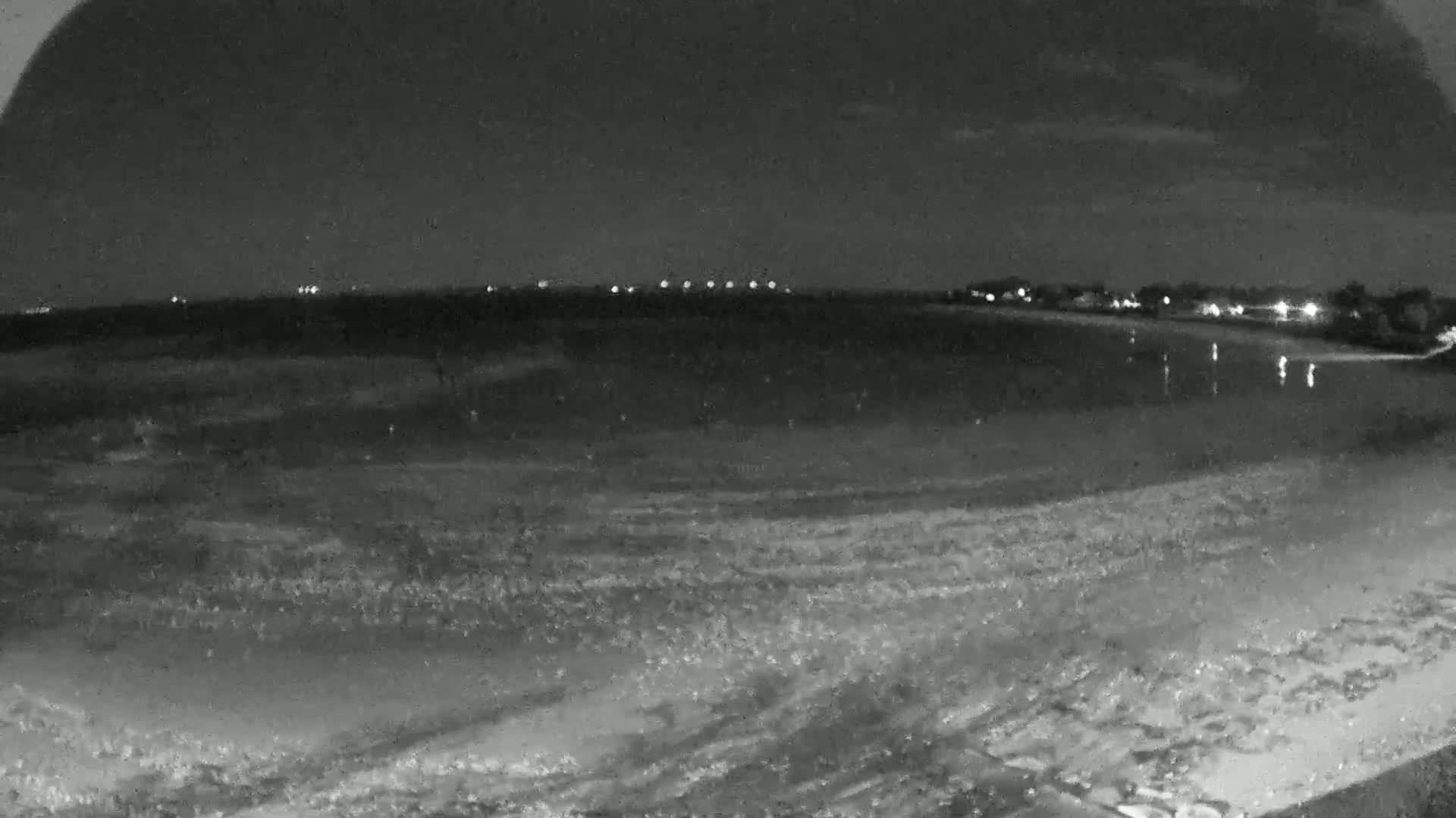 A nighttime beach scene shows gently lapping waves on a sandy shore under a mostly clear sky, with lights visible in the distance across the water.