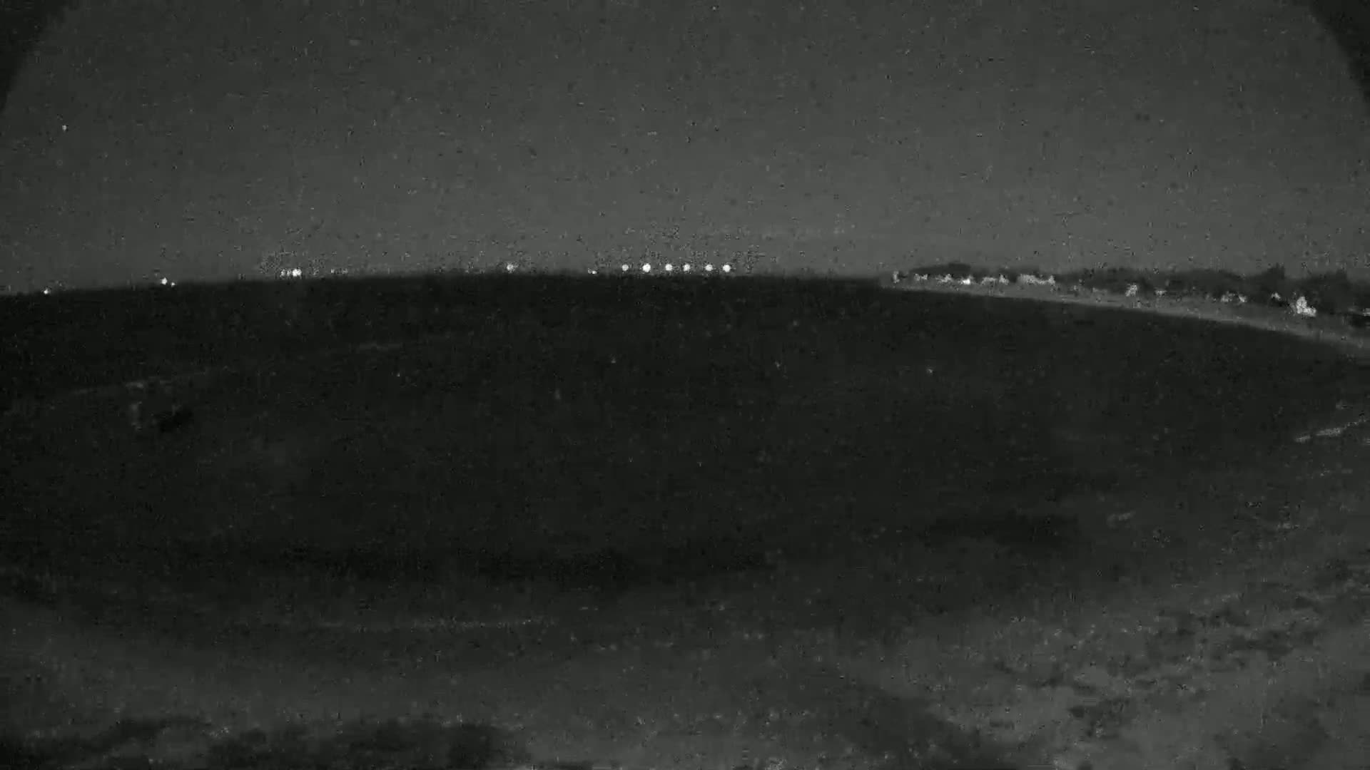 A dark, nighttime landscape shows a line of distant lights on the horizon under a mostly clear sky.
