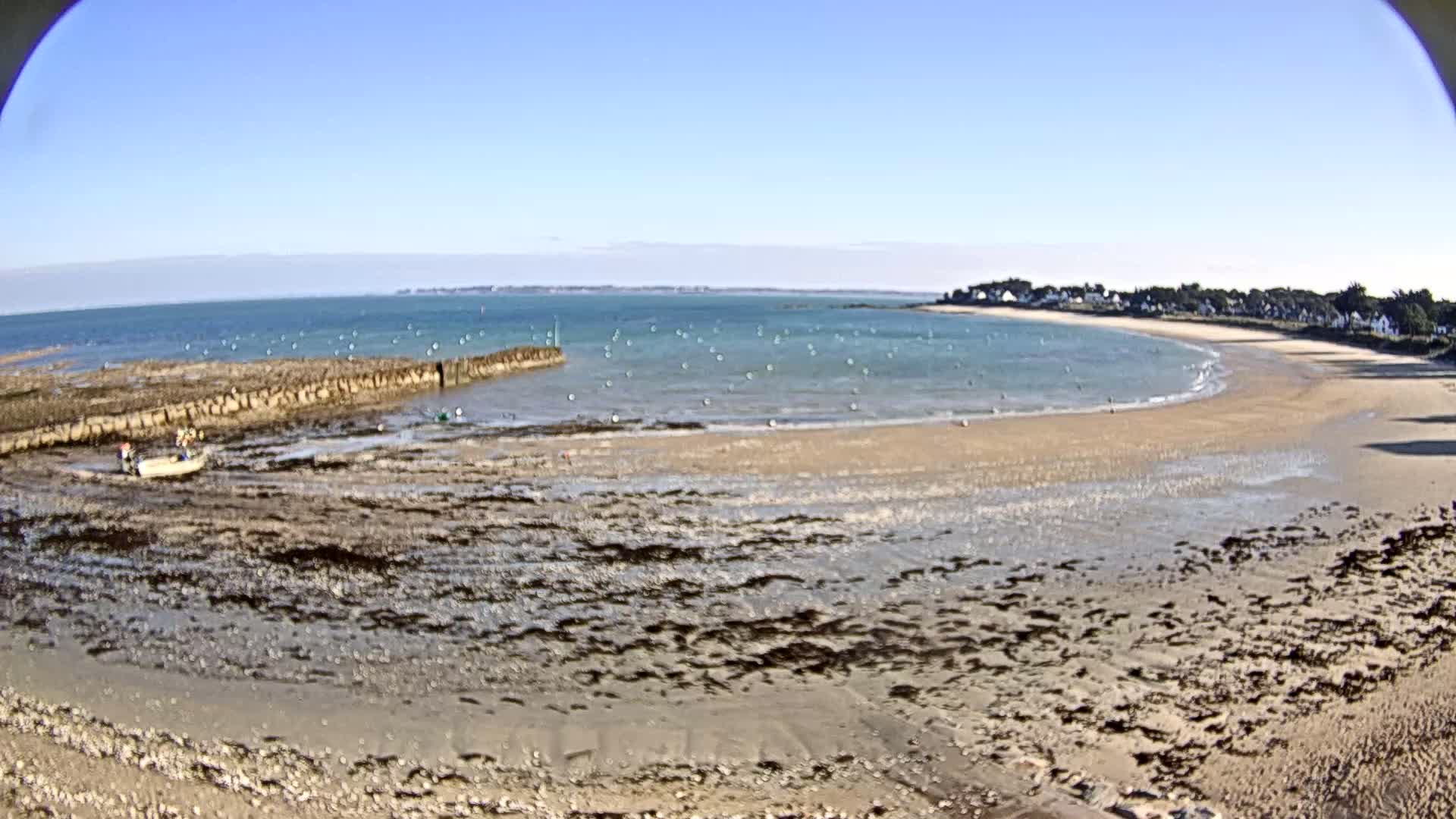 A sandy beach curves around a calm bay under a clear blue sky, with a small boat near a rock jetty and a few people visible on the shore.