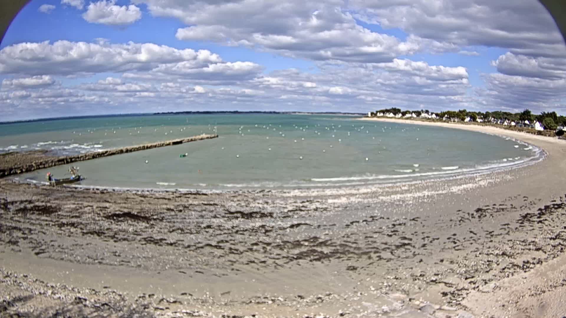 A partly cloudy day reveals a sandy beach curving around a calm bay dotted with small boats, a small fishing boat near a jetty, and a small village in the distance.