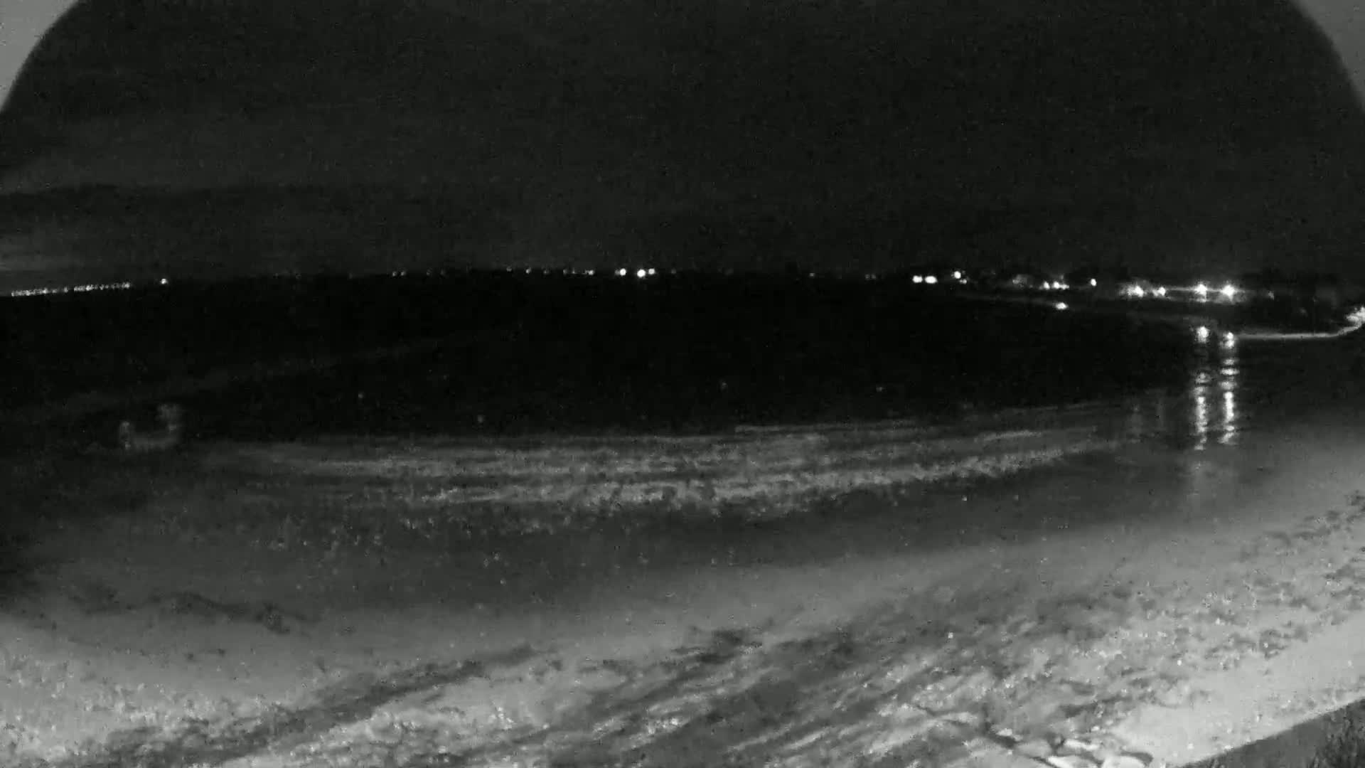 A nighttime view of a dark beach with gently lapping waves and distant lights reflecting on the water.