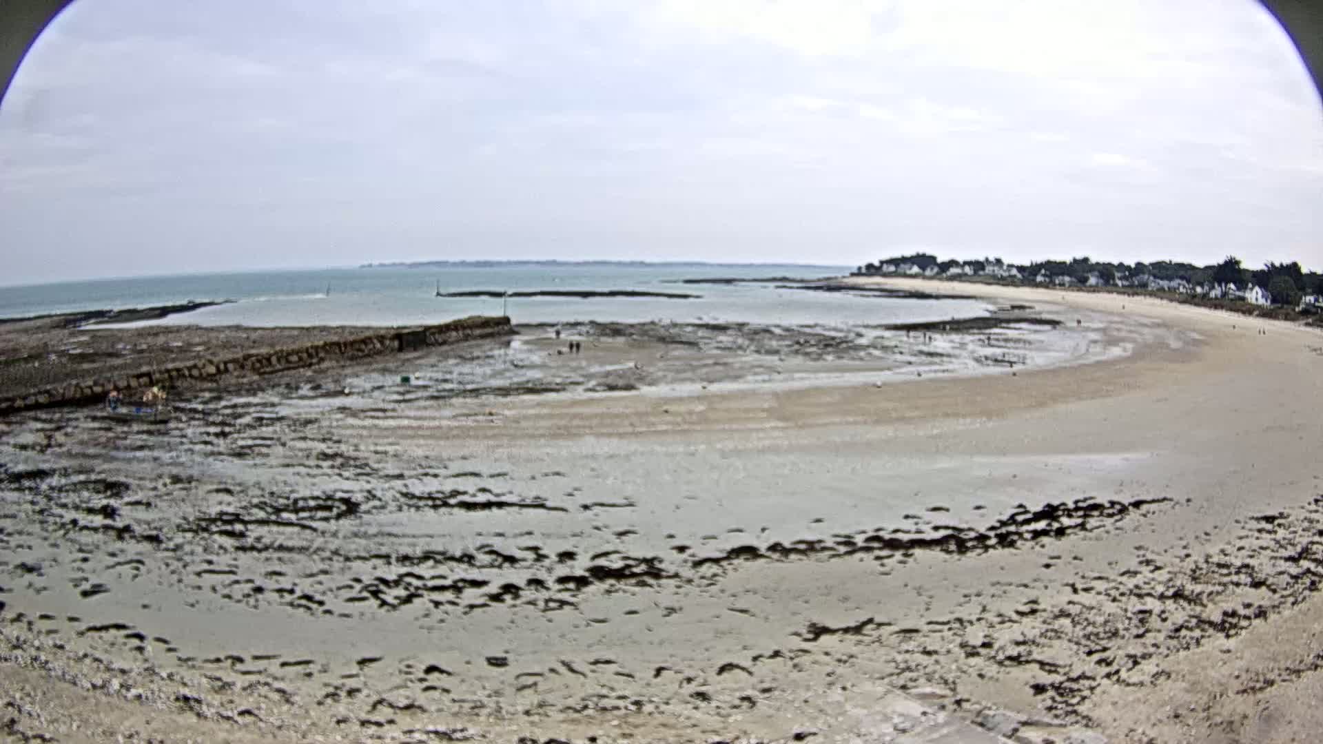 A wide shot shows a mostly sandy beach at low tide, with a calm sea and a small village visible in the distance under a cloudy sky.