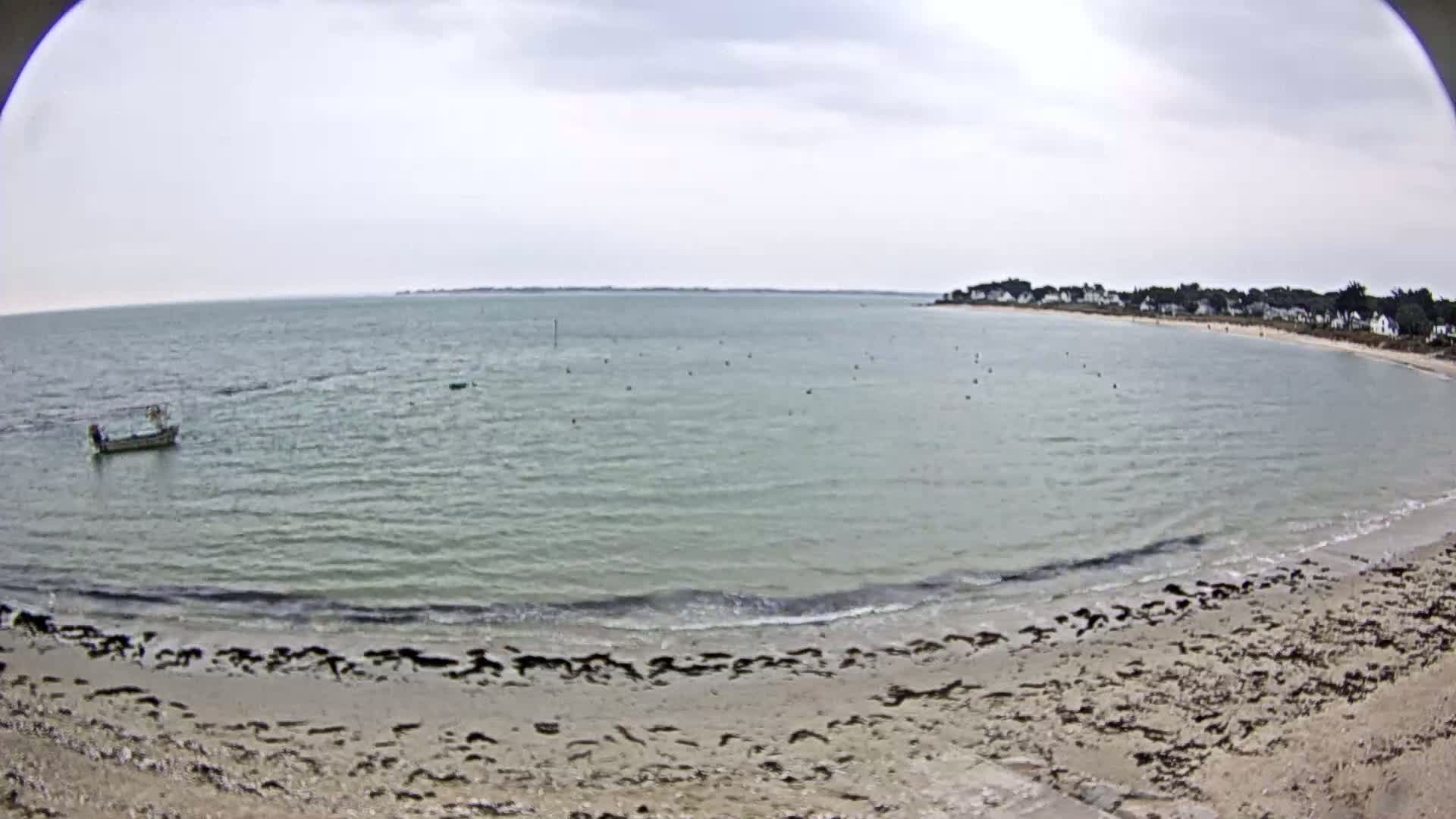A sandy beach with a calm, grey-green sea under an overcast sky; a small boat is visible in the water.