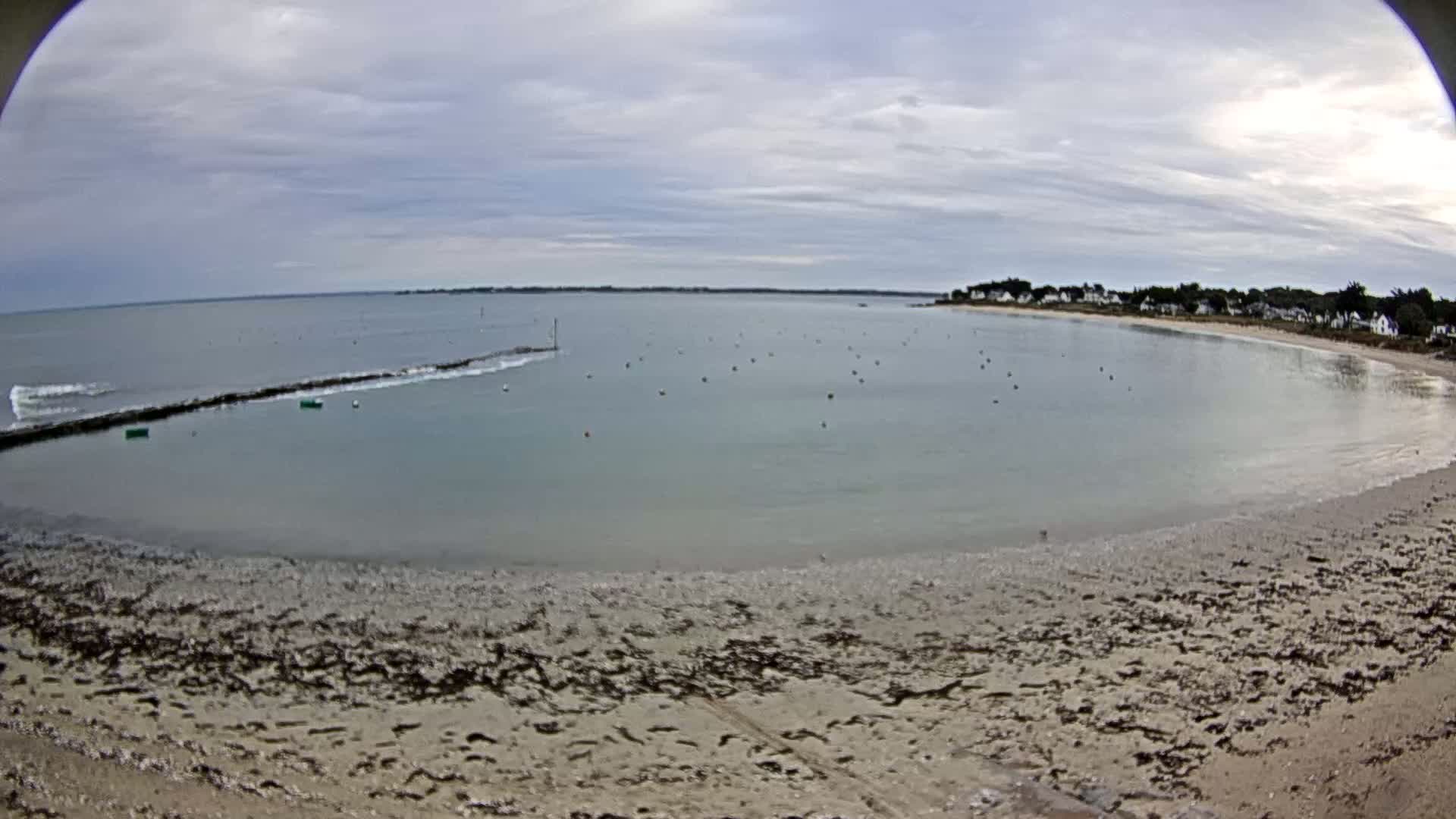 A calm, overcast day at a sandy beach with a small town visible in the distance, showing a breakwater and some buoys in the shallow water.