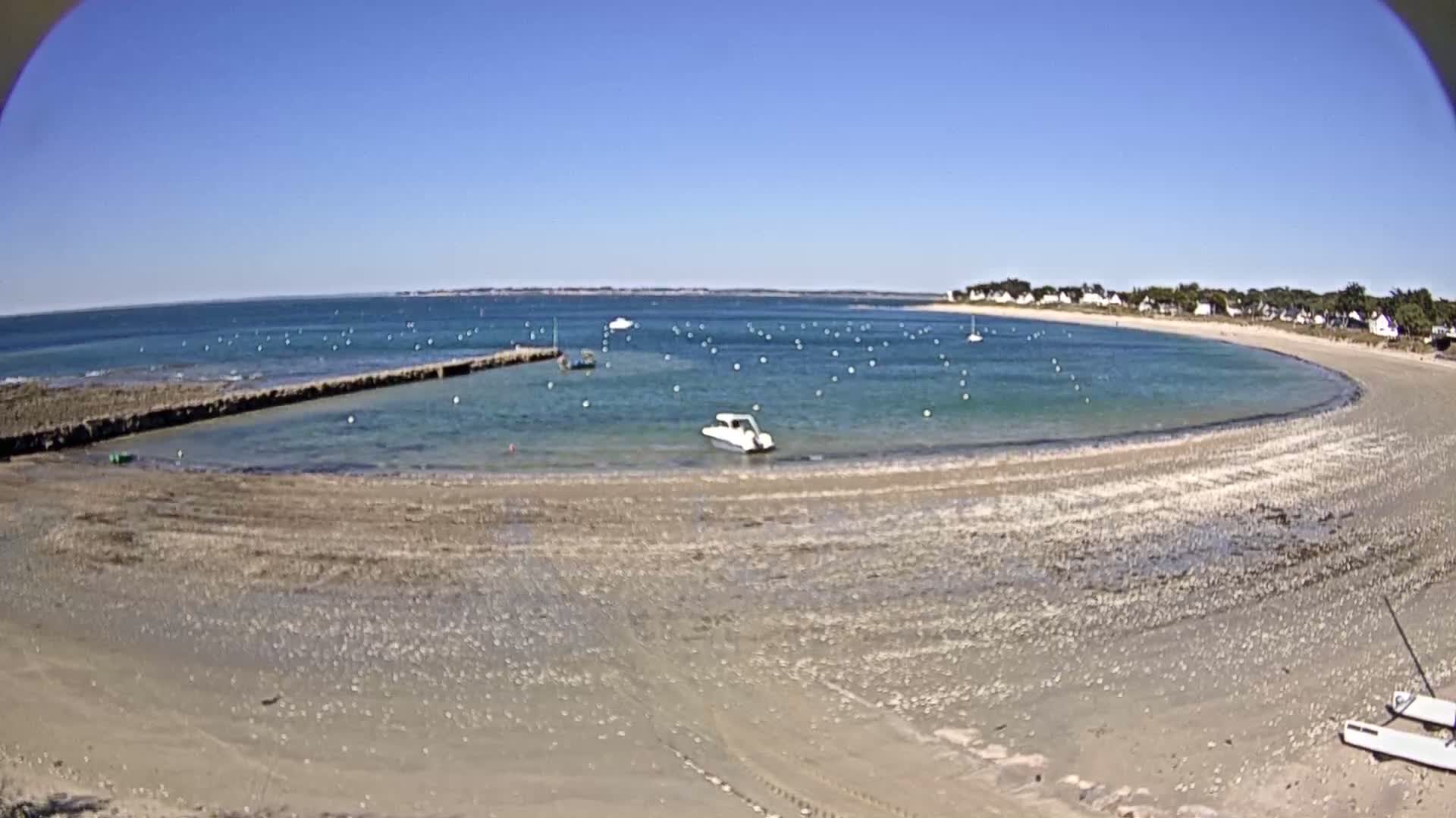 A sandy beach curves around a calm, clear bay dotted with small boats and buoys under a bright, sunny sky.