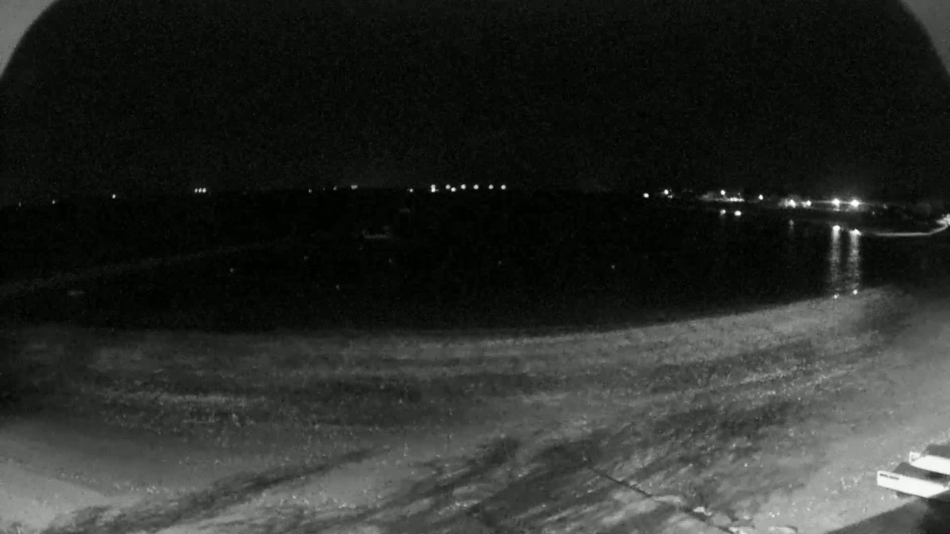 A nighttime, low-light view of a dark body of water with a shoreline showing some texture, and distant lights visible across the water.