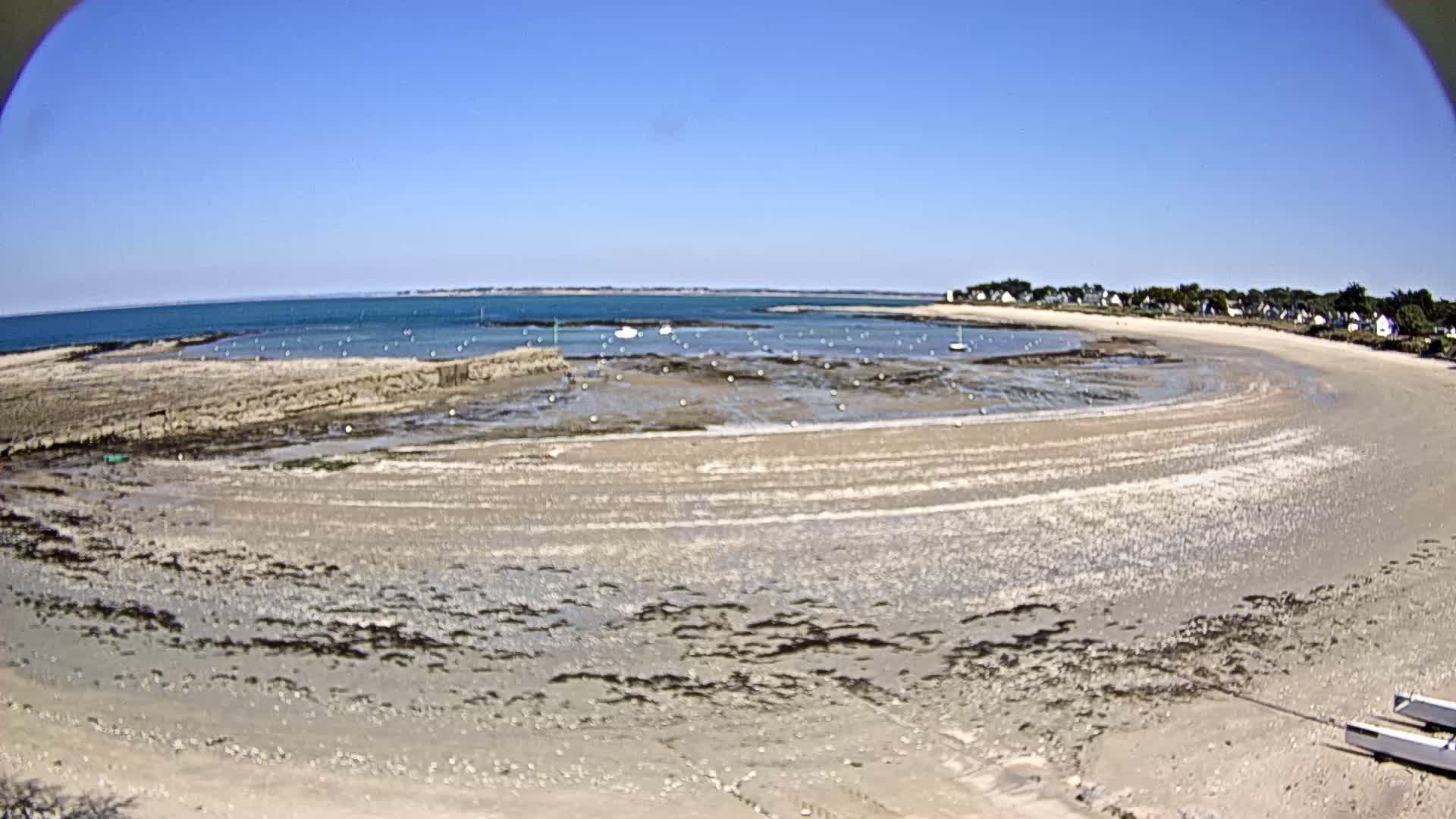 A sandy beach curves around a calm bay under a clear blue sky, with several small boats visible in the shallow water near the shore.