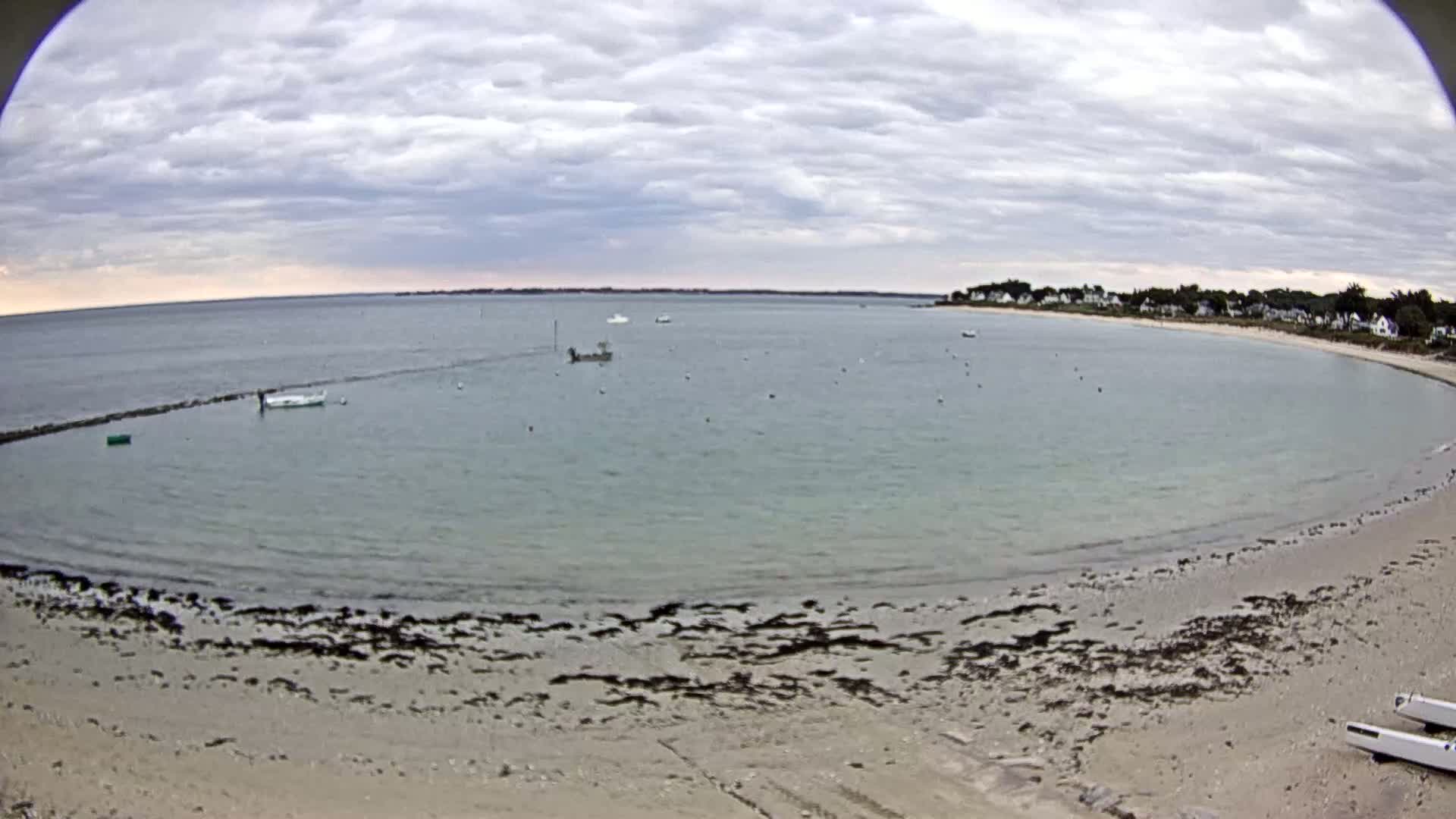 A sandy beach curves around a calm bay under a cloudy sky, with several small boats visible in the water and houses on the far shore.
