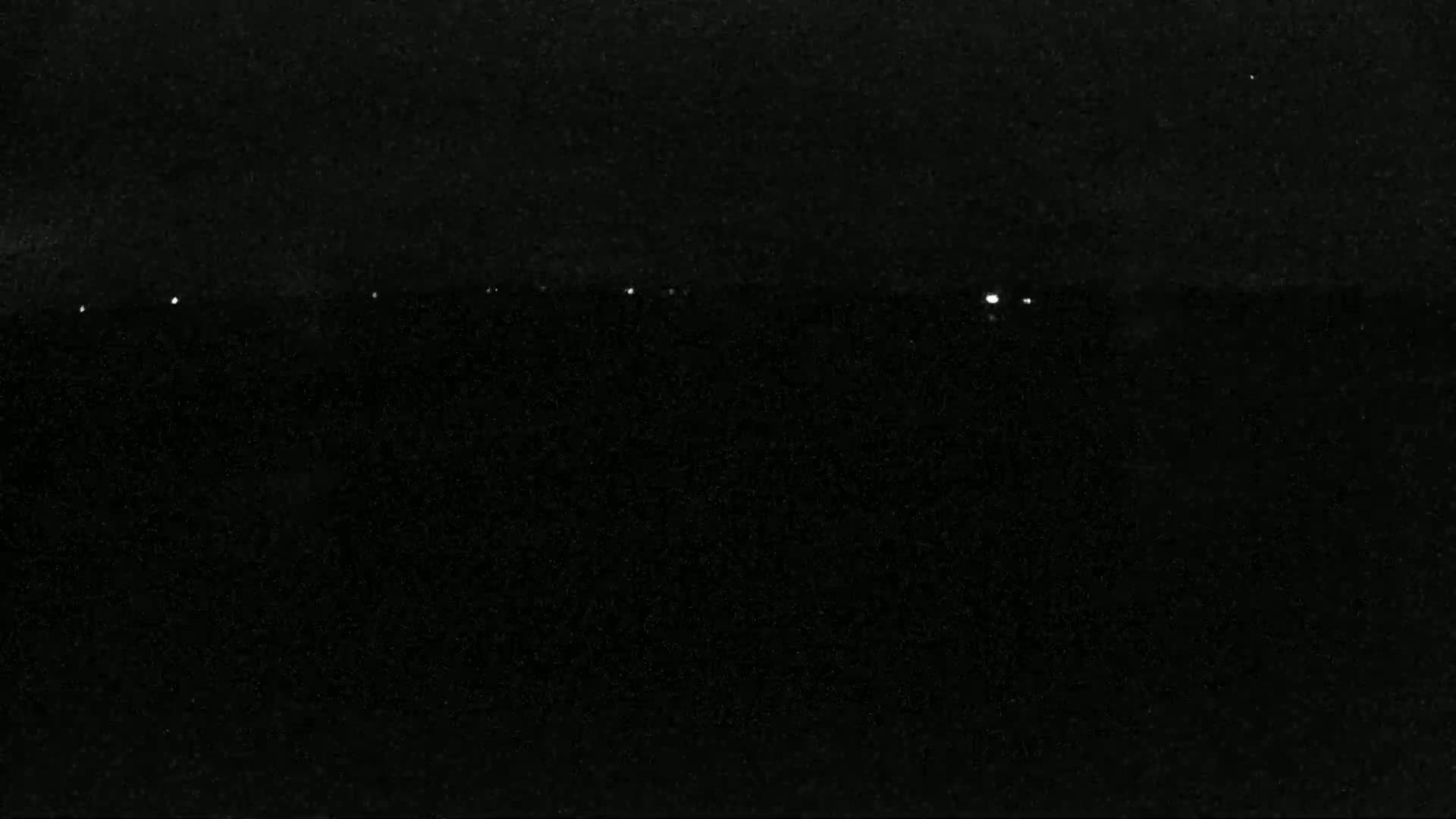 A nighttime, low-light image shows a dark expanse with a few faint, distant lights on the horizon.