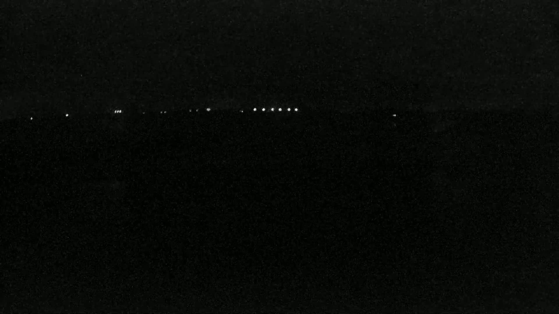 A nighttime, dark landscape is illuminated by a distant line of lights.