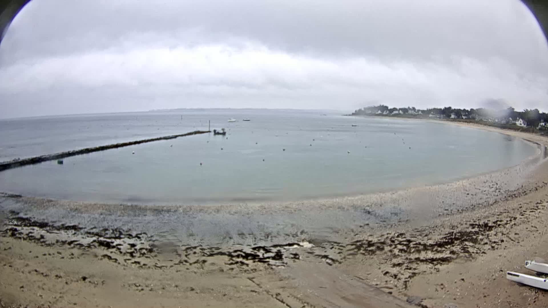 A calm, overcast day reveals a sandy beach curving into a sheltered bay with a few boats and a stone jetty, under a mostly cloudy sky.