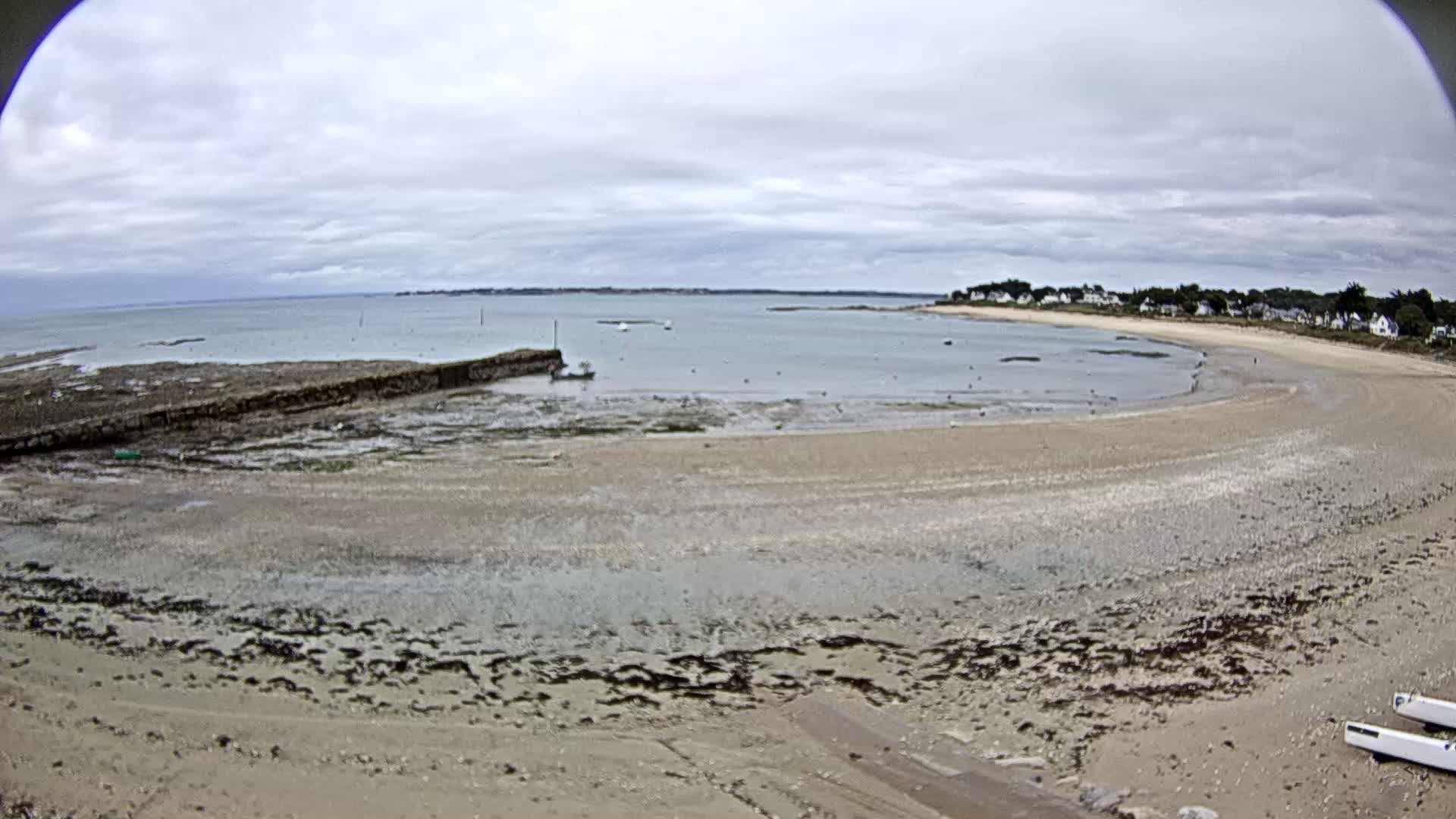 A wide, sandy beach curves around a calm bay under a cloudy sky, with a few boats visible in the water and a low stone wall near the shore.