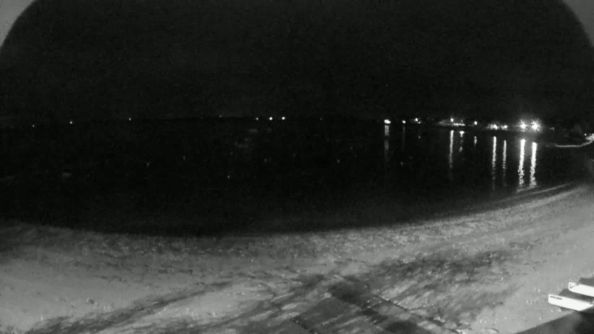 A dark, nighttime beach scene shows a calm body of water reflecting distant lights, with a sandy shore in the foreground.
