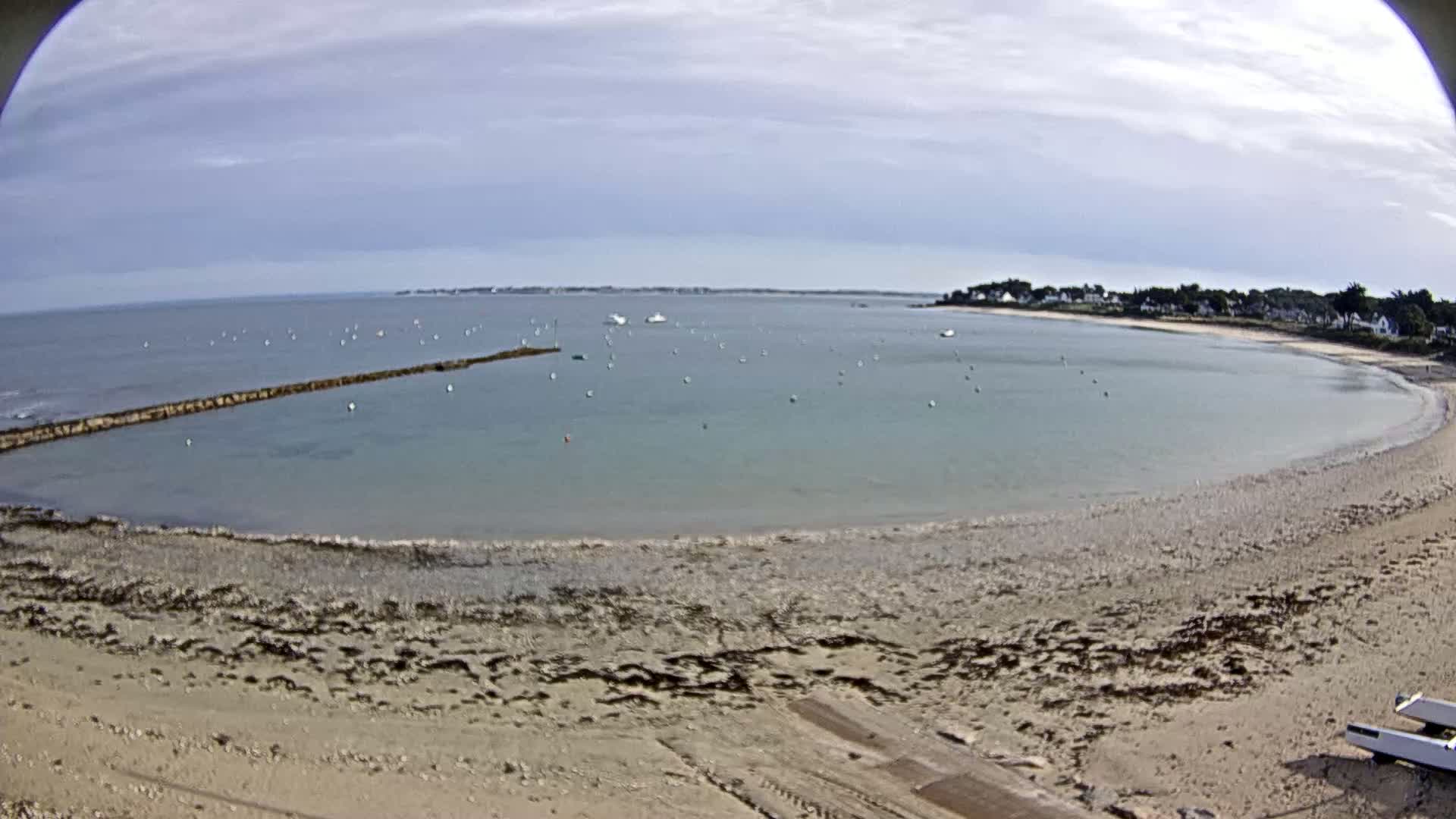 A sandy beach curves around a calm, light-blue bay dotted with numerous small boats under a mostly cloudy sky.