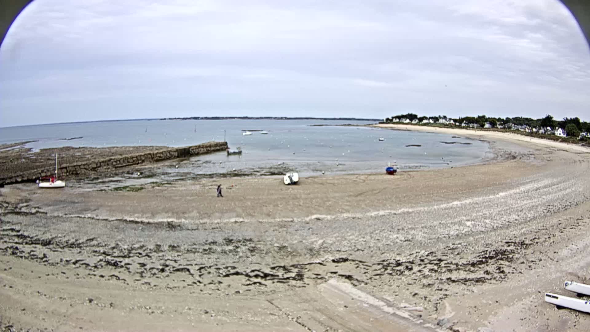 A mostly sunny day reveals a sandy beach and calm bay with several small boats, a low stone wall, and a few people walking near the water's edge.
