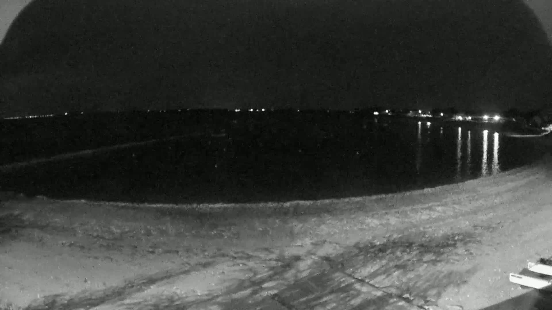 A dark, nighttime view of a calm body of water reflecting distant lights on a sandy shoreline.