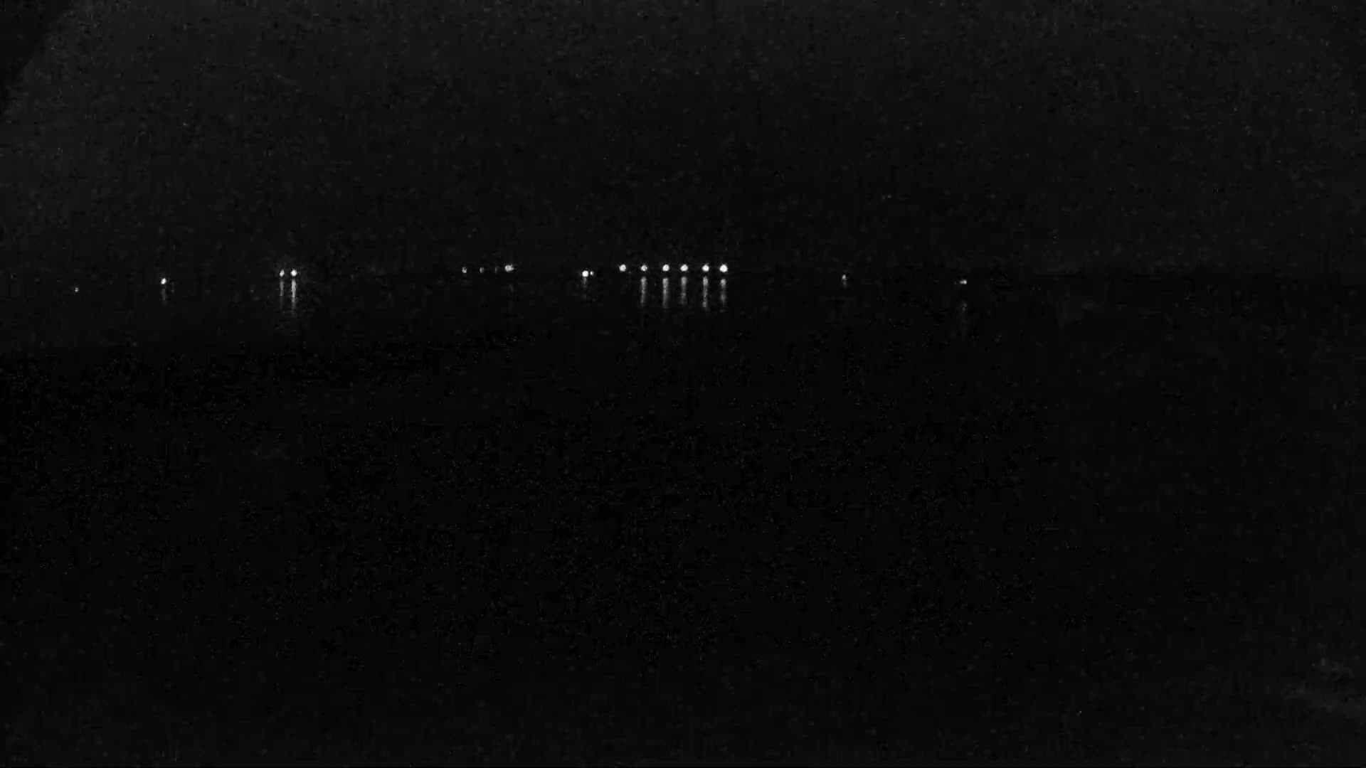 A dark, nighttime scene shows a body of water reflecting a few distant lights.