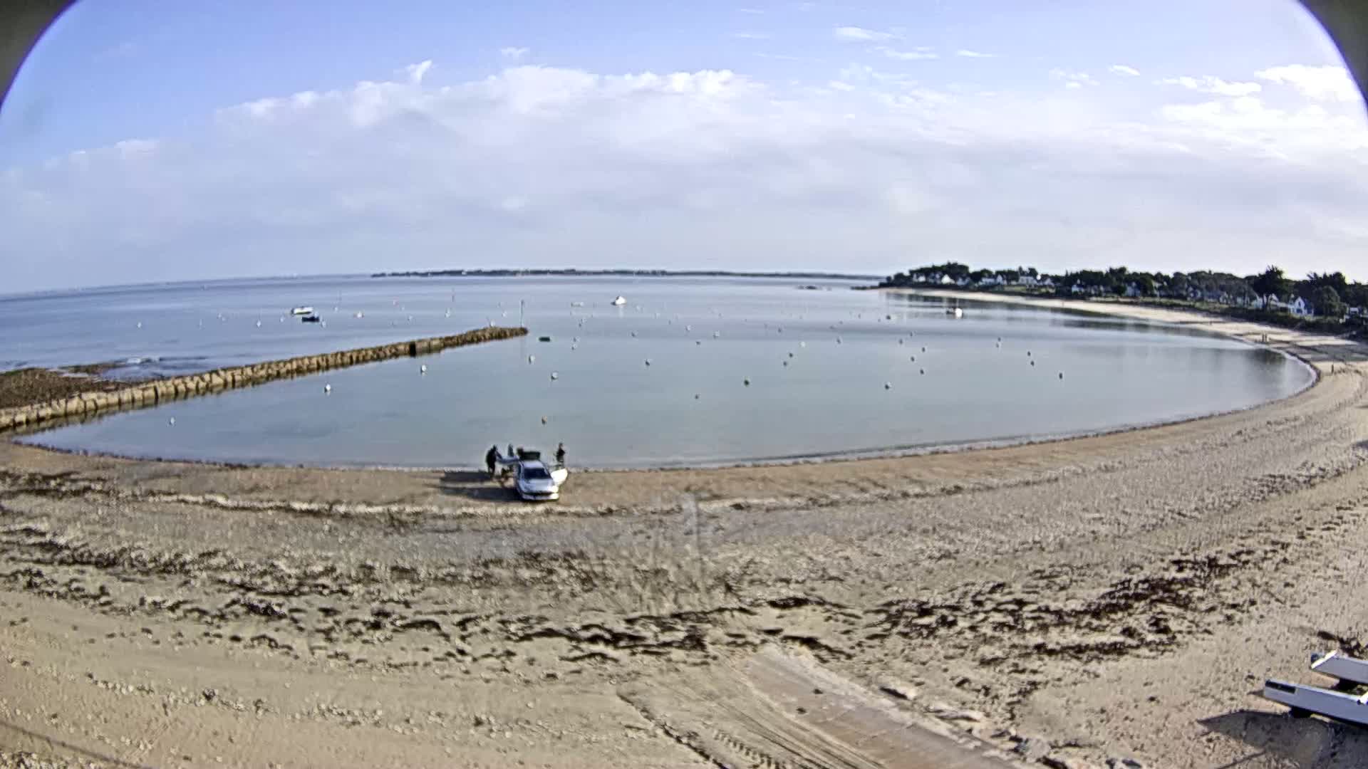 A sandy beach curves around a calm bay under a partly cloudy sky, with a car and several people near the water's edge and a few boats in the distance.