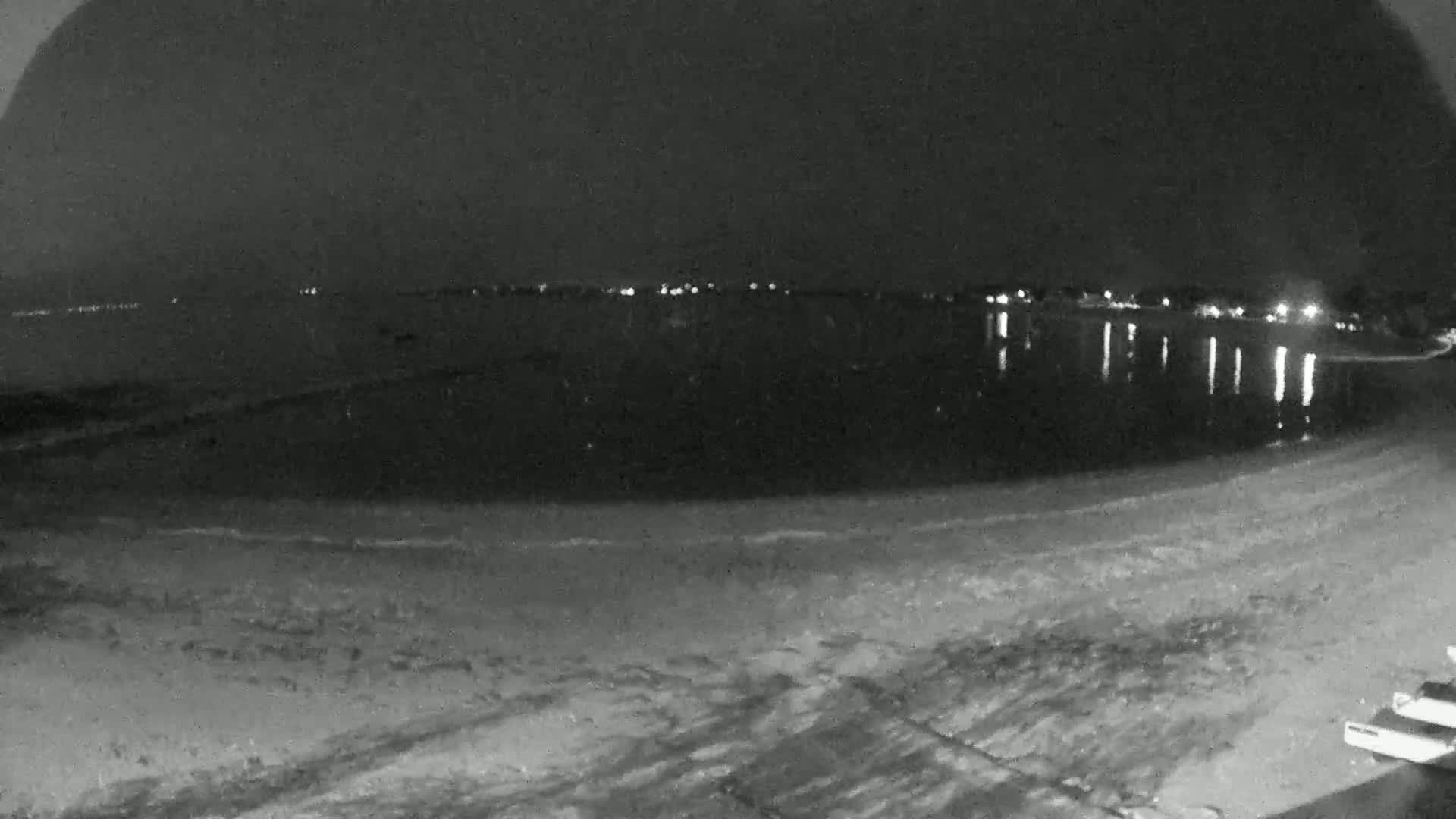 A dark, nighttime beach scene shows calm water reflecting distant lights, with a mostly empty sandy shore in the foreground.