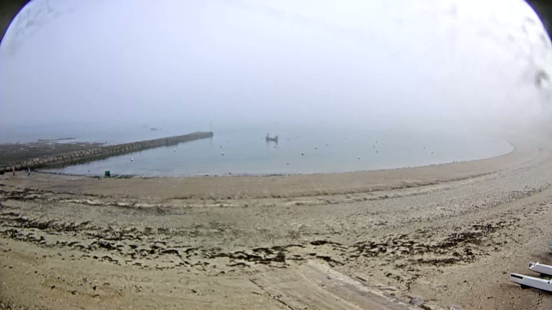 A sandy beach curves around a calm, foggy bay with a stone breakwater and a few small boats visible in the distance.