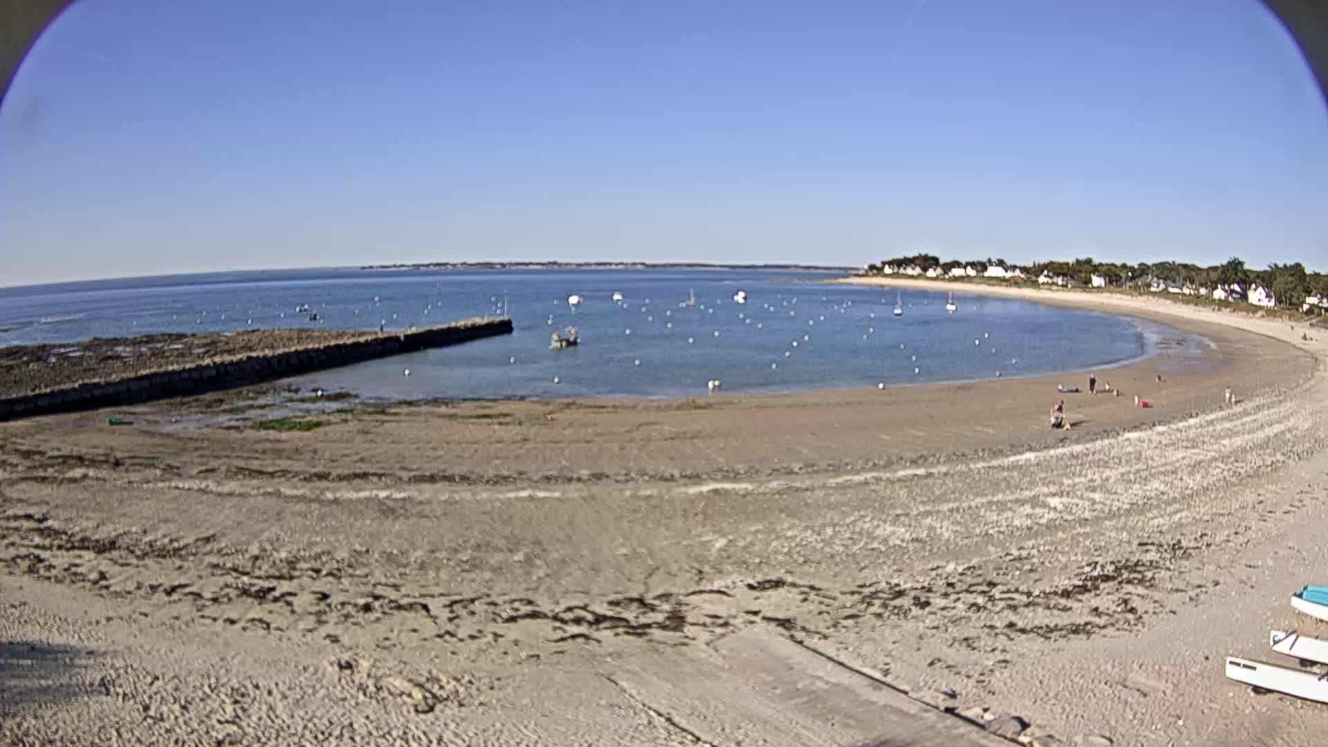 A sandy beach curves around a calm bay dotted with small boats under a clear blue sky.