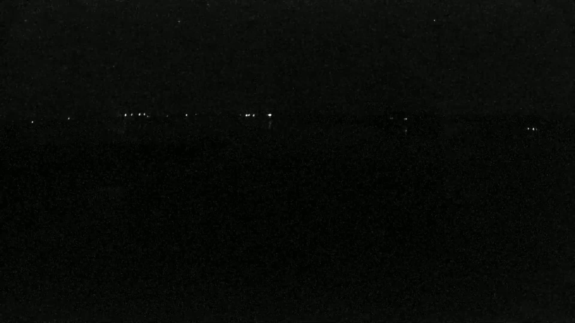 A dark, nighttime scene shows a distant line of faint lights across a mostly black expanse.